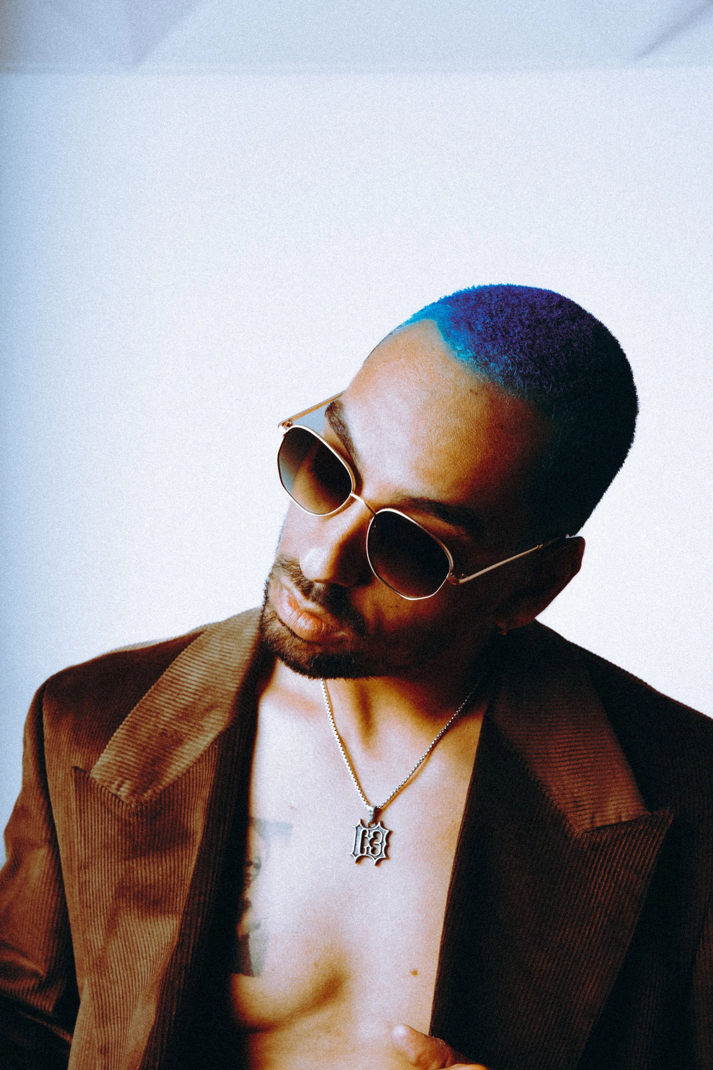 A man with a blue and black hairstyle wearing sunglasses and a brown blazer, showing part of his chest with a tattoo and a necklace with a letter 'B' pendant.