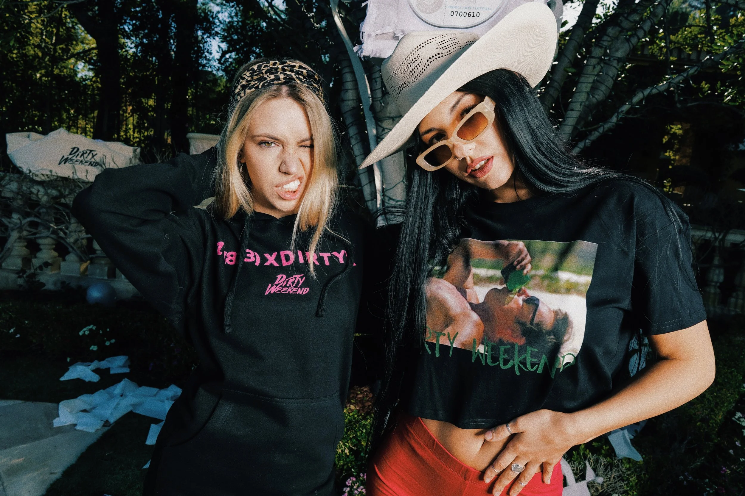 Two women posing outdoors for a photo, wearing casual black clothing with 'Dirty Weekend' text, one with a wide-brimmed hat and sunglasses, the other with a leopard print headband. They are making expressive faces and are standing close to each other