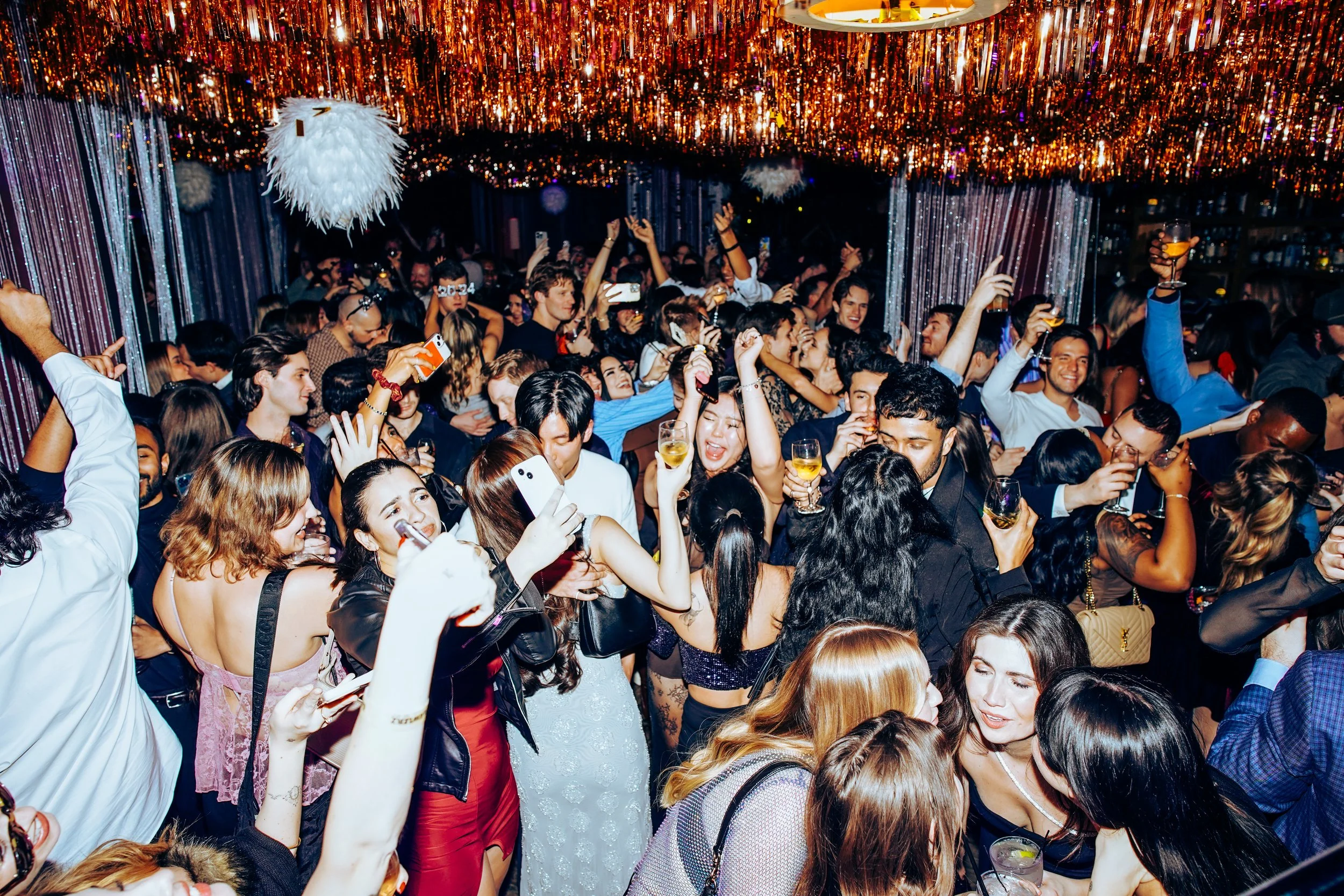 A crowded party with people dancing, taking photos, and holding drinks under colorful decorations and lights.
