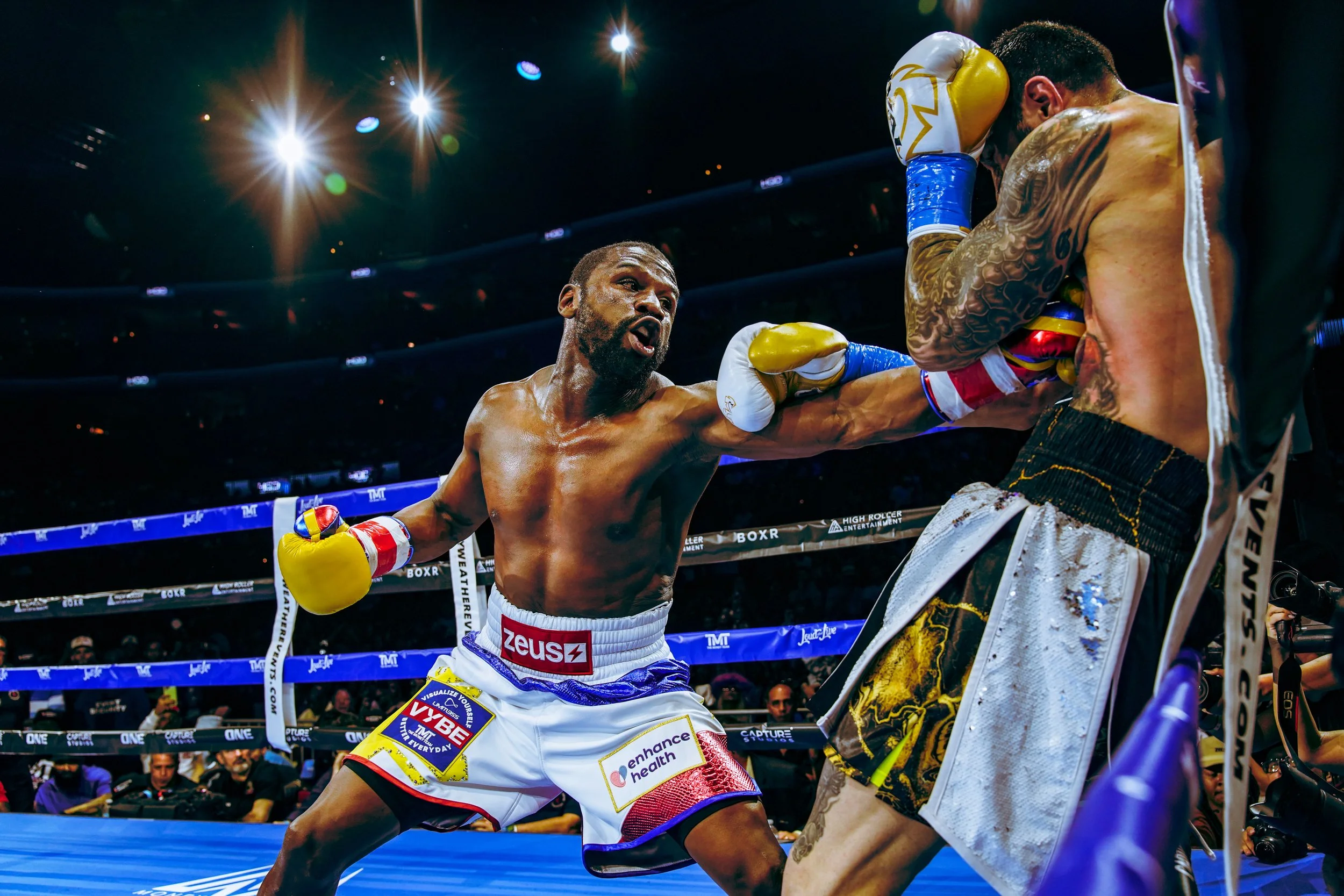 Two boxers in a boxing ring during a match, one throwing a punch at the other, who is covering up. Bright lights overhead and audience in the background.