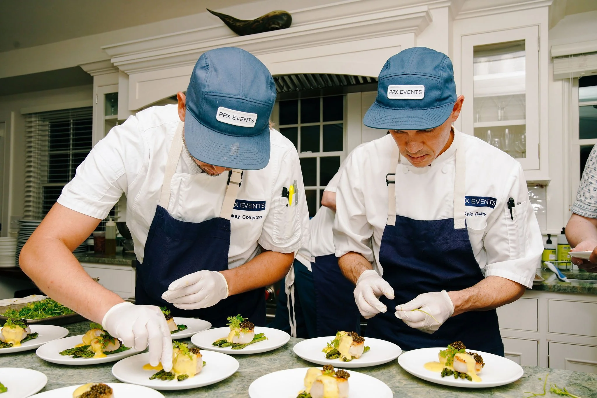 Two chefs in white uniforms and blue caps preparing gourmet dishes in a professional kitchen.