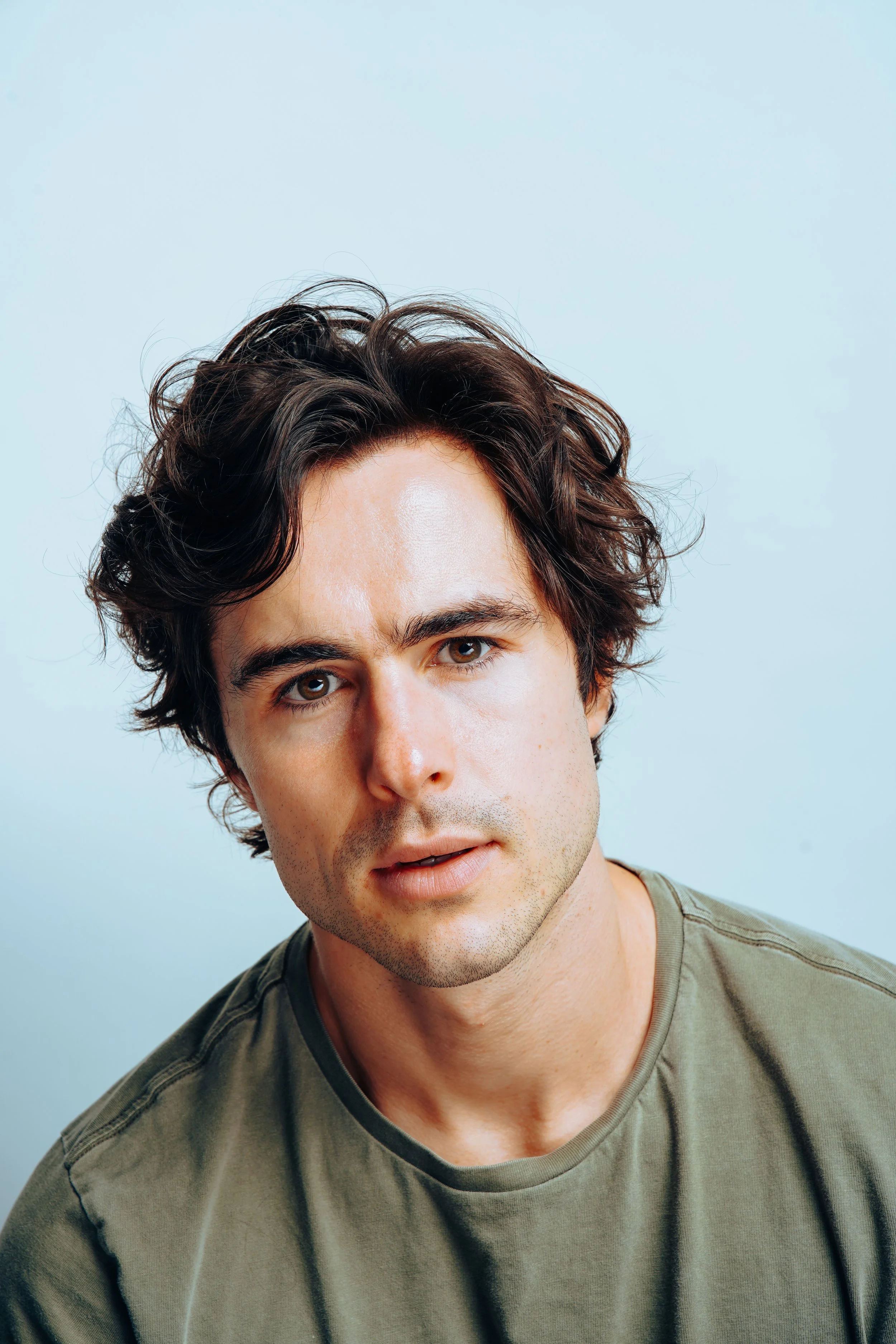 Close-up of a young man with dark wavy hair wearing a green t-shirt, looking directly at the camera with a neutral expression against a light blue background.