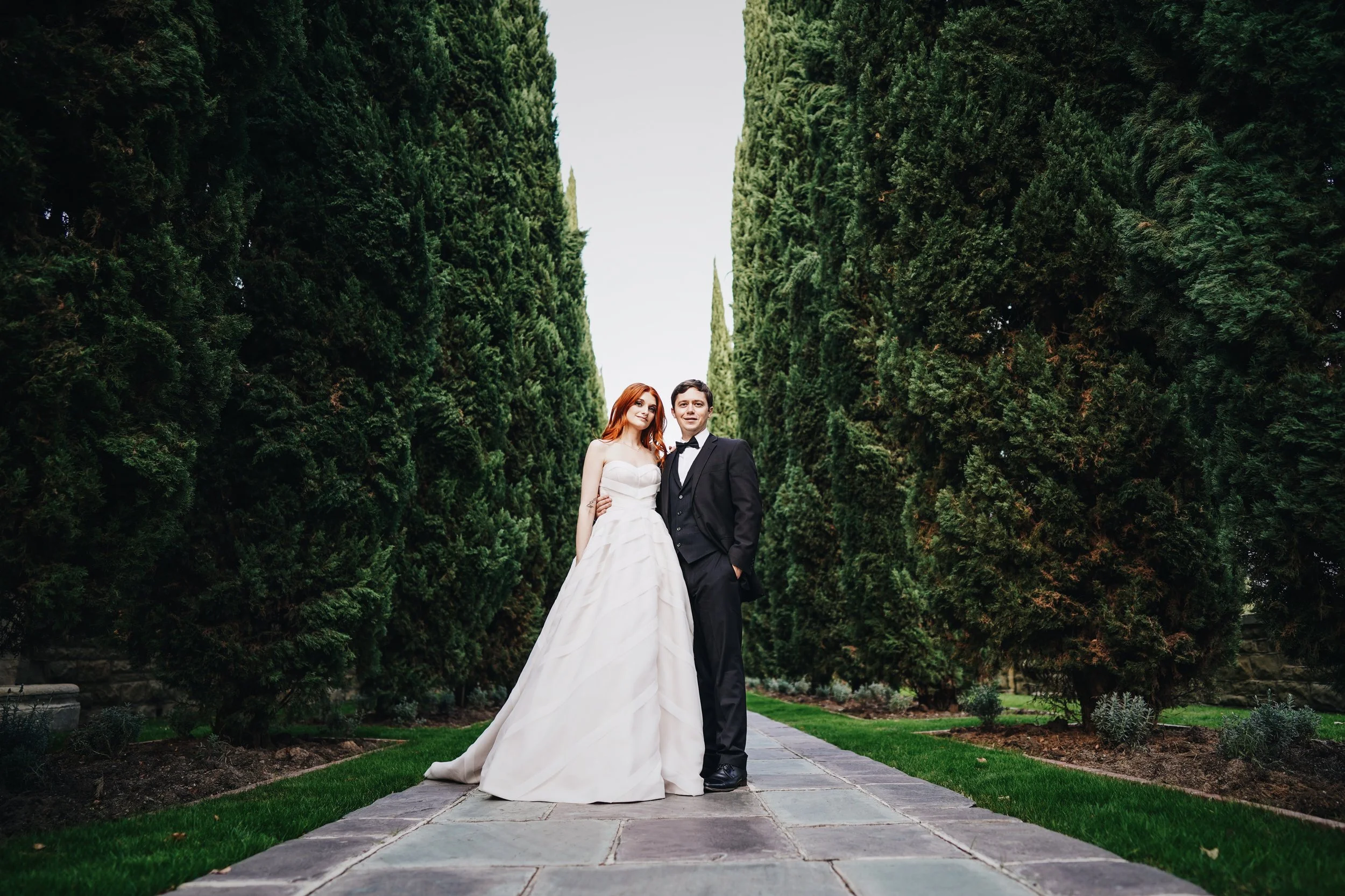 A bride and groom standing together on a stone pathway, surrounded by tall green trees, during daytime.