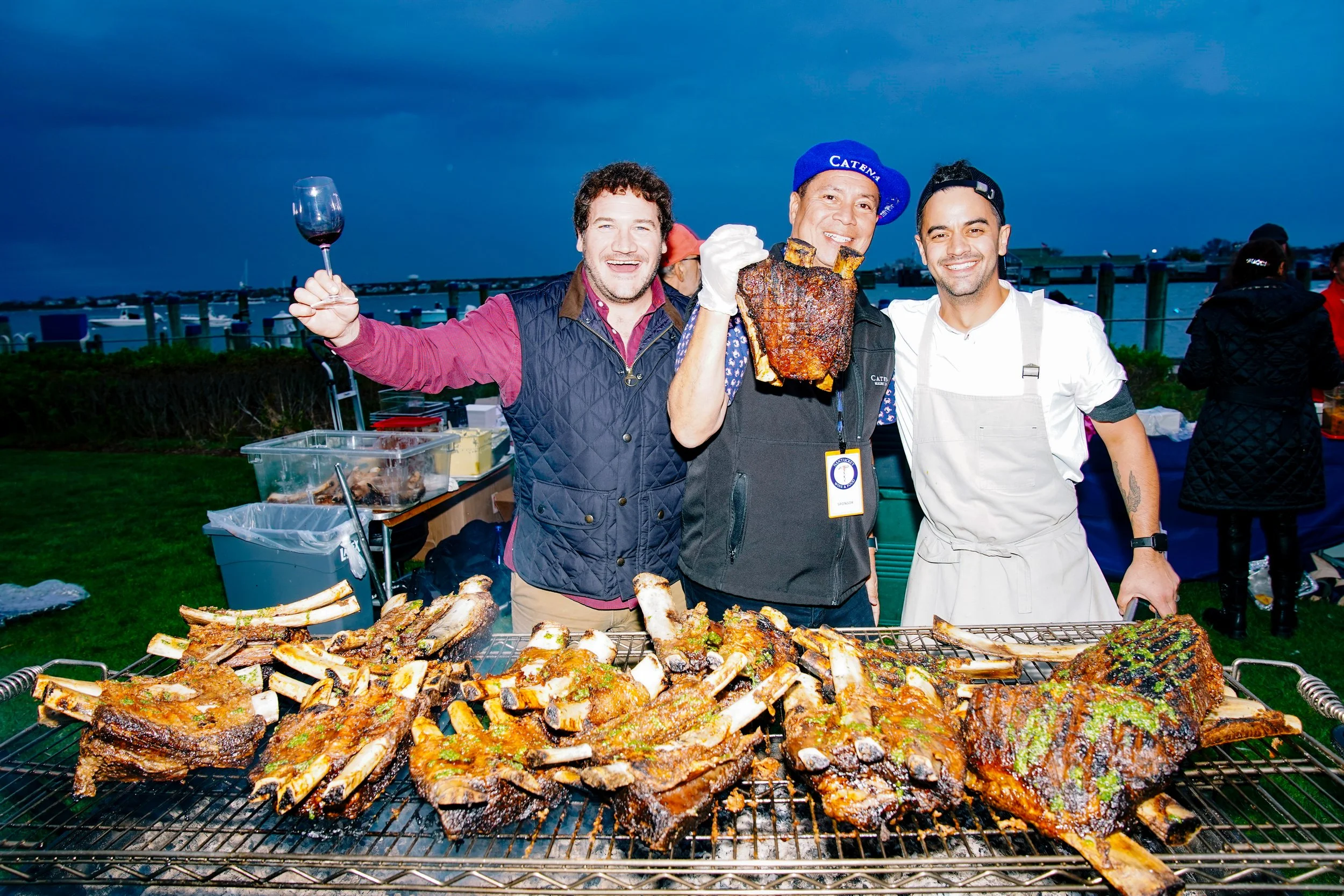 Three men at an outdoor barbecue by the water, smiling and holding up grilled meats, with ribs on the grill in front of them.