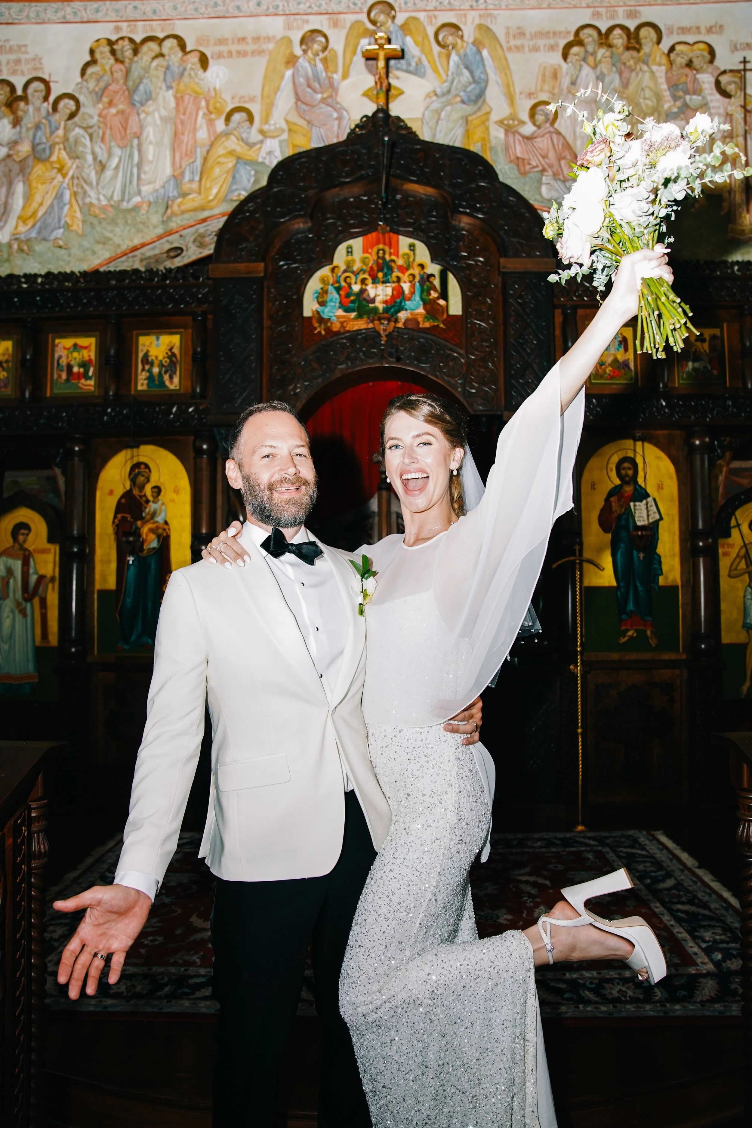 A bride and groom celebrating their wedding in a church with religious icons and paintings in the background. The bride has one arm raised with a bouquet of flowers, and both are smiling.