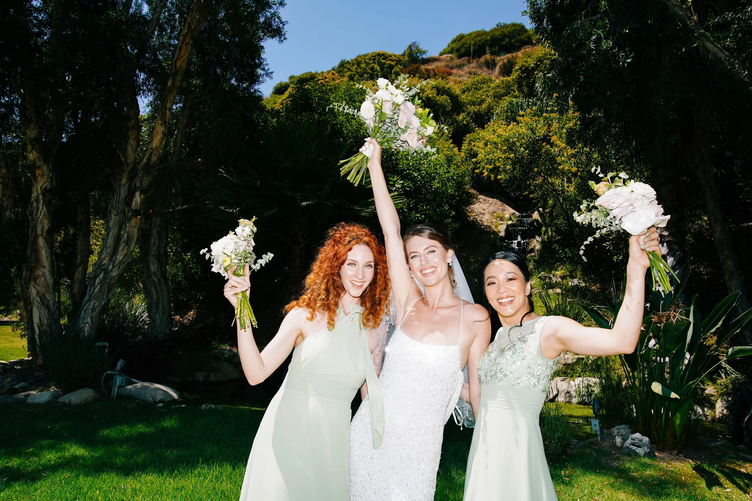 Three women in dresses celebrating outdoors, holding bouquets, with one woman raising her bouquet in the air, surrounded by trees and greenery.