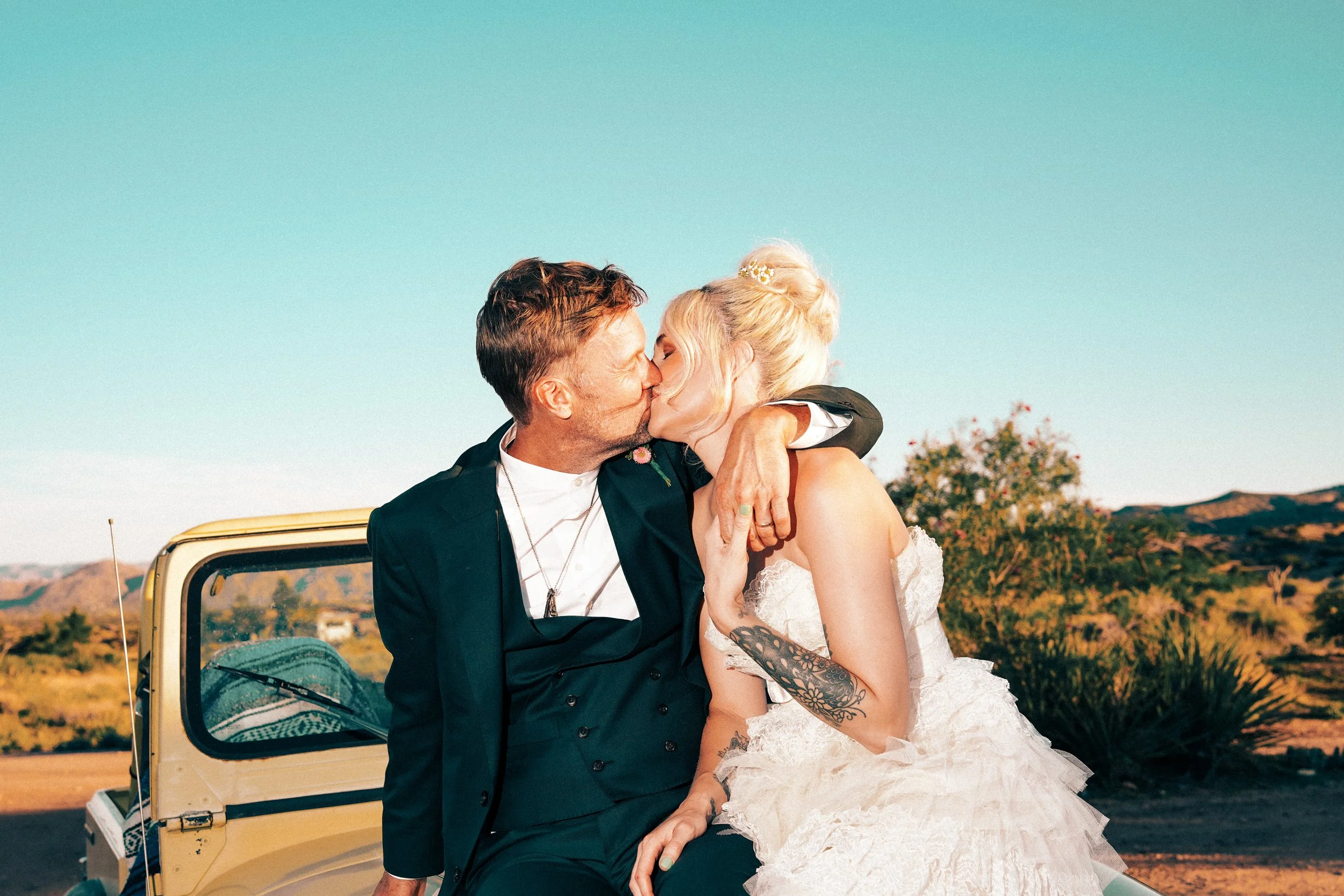 A man and woman in wedding attire share a kiss outdoors with a yellow vehicle and desert landscape in the background.