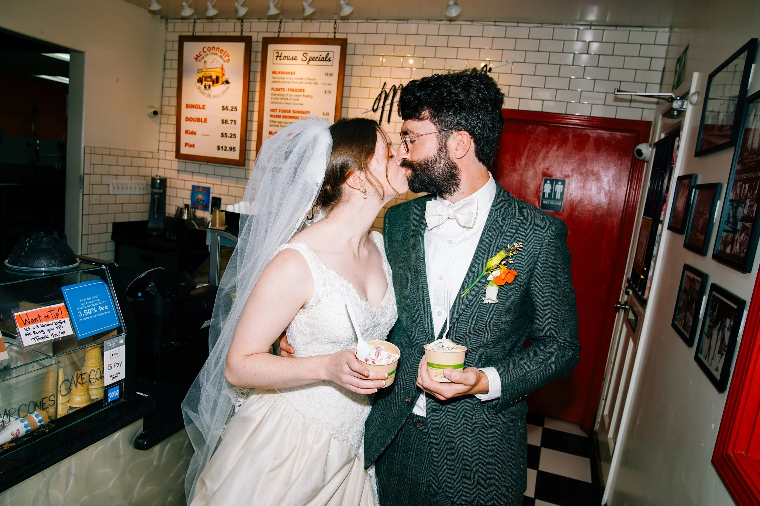 A bride and groom sharing a kiss inside an ice cream shop, holding cups of ice cream. The bride is wearing a wedding dress and veil, and the groom is wearing a grey suit with a bow tie and boutonniere. There are menus and framed pictures on the wall 