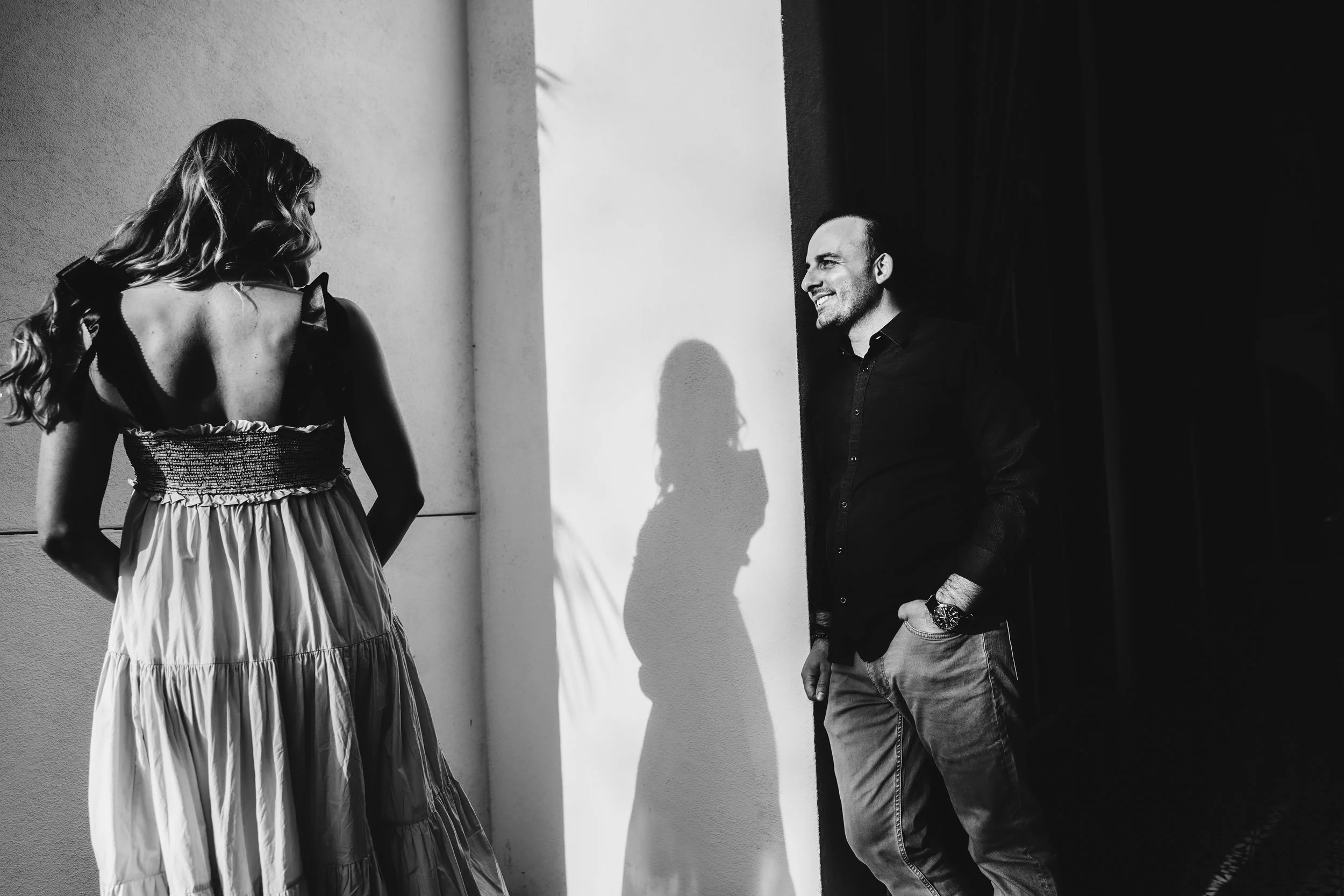 A woman in a strapless dress faces a man in dark clothing, both smiling, with their shadows cast on the wall between them.
