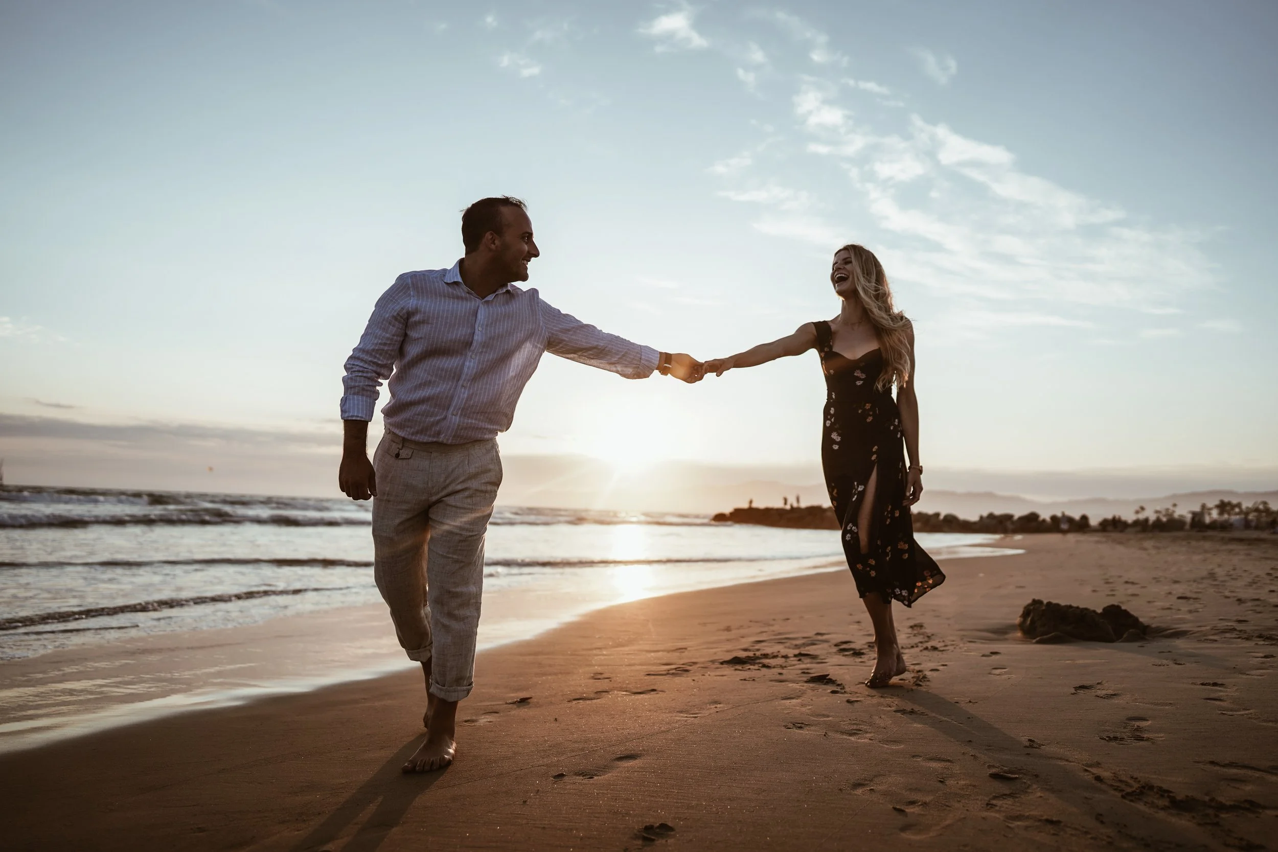 A couple holding hands and walking on the beach at sunset, smiling and enjoying each other's company.