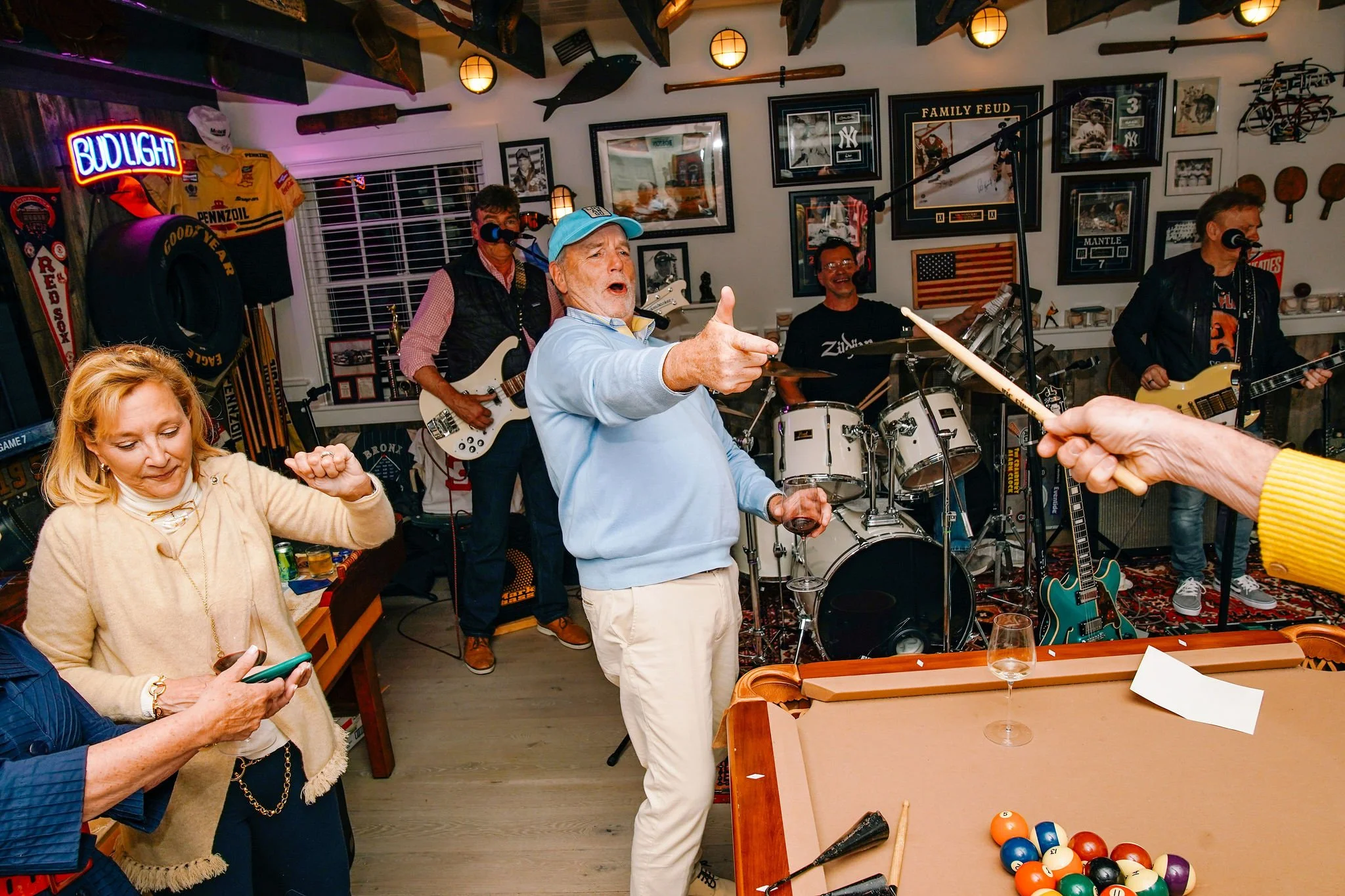 A lively indoor gathering with a band performing in a decorated room, a woman dancing, an older man with a drink singing with encouragement, people taking selfies, pool table in foreground, and various sports memorabilia and pictures on the walls.