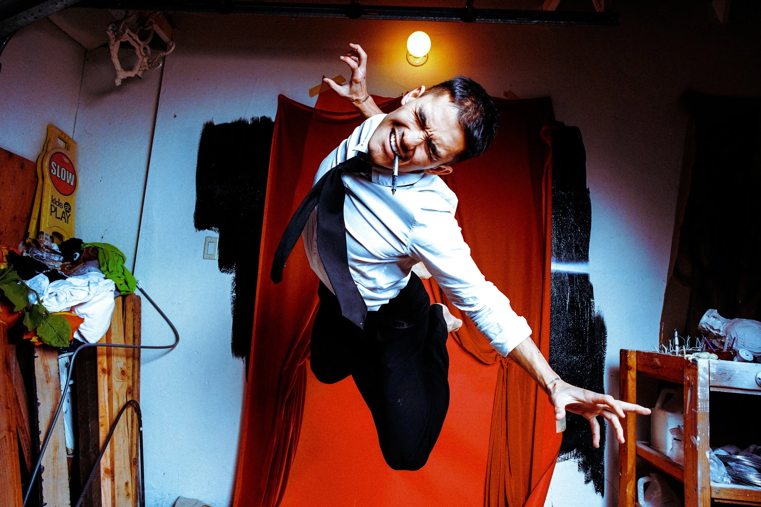 A man in a white shirt, black pants, and a black tie is posing dynamically, leaning forward with one hand on a wooden surface and a pen in his mouth, in front of a red backdrop in a cluttered room.