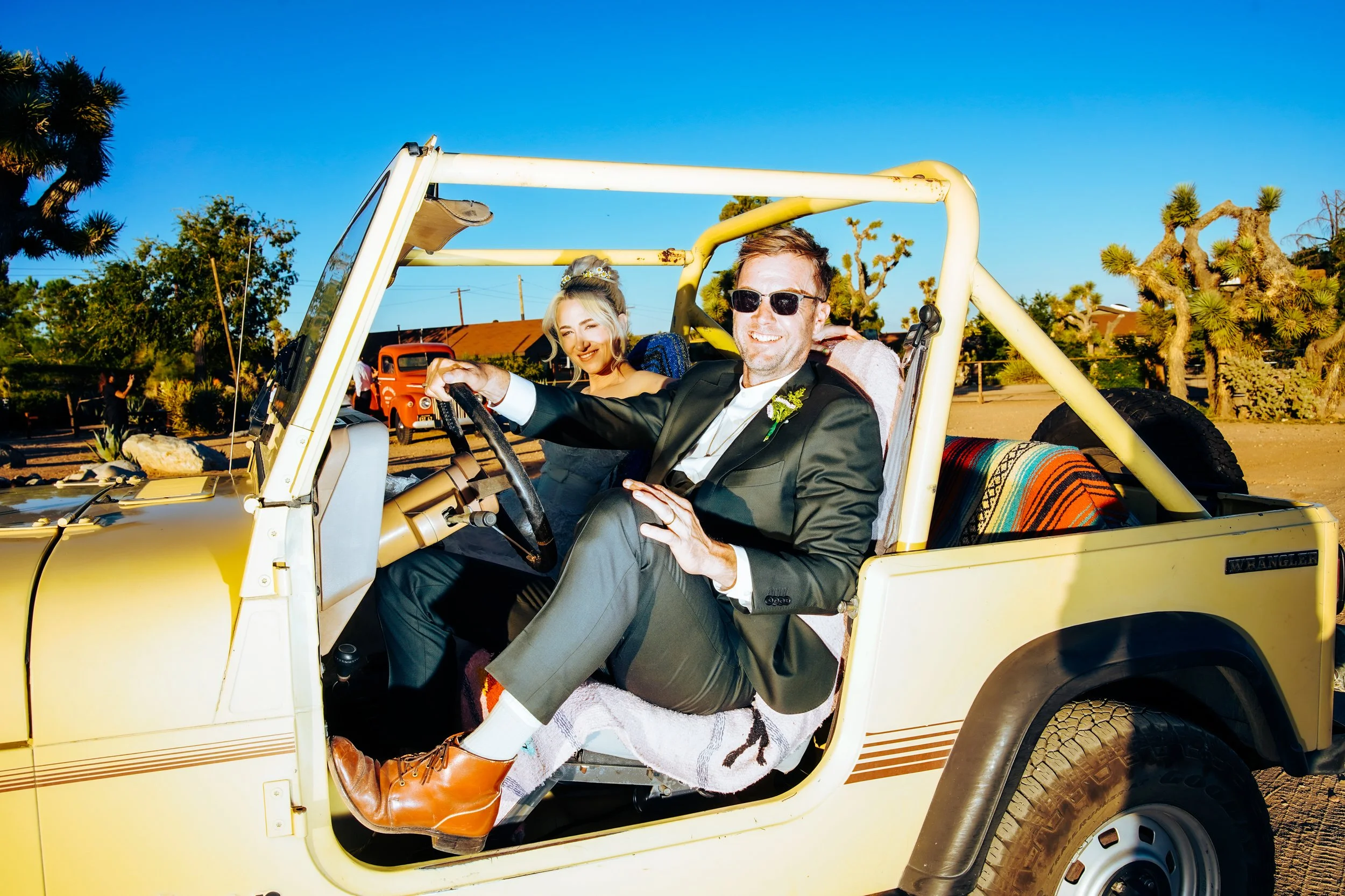 Two people sitting in a yellow Jeep, a man in a suit with sunglasses smiling and a woman with blonde hair smiling, in a desert-like setting with Joshua trees and old vehicles in the background.