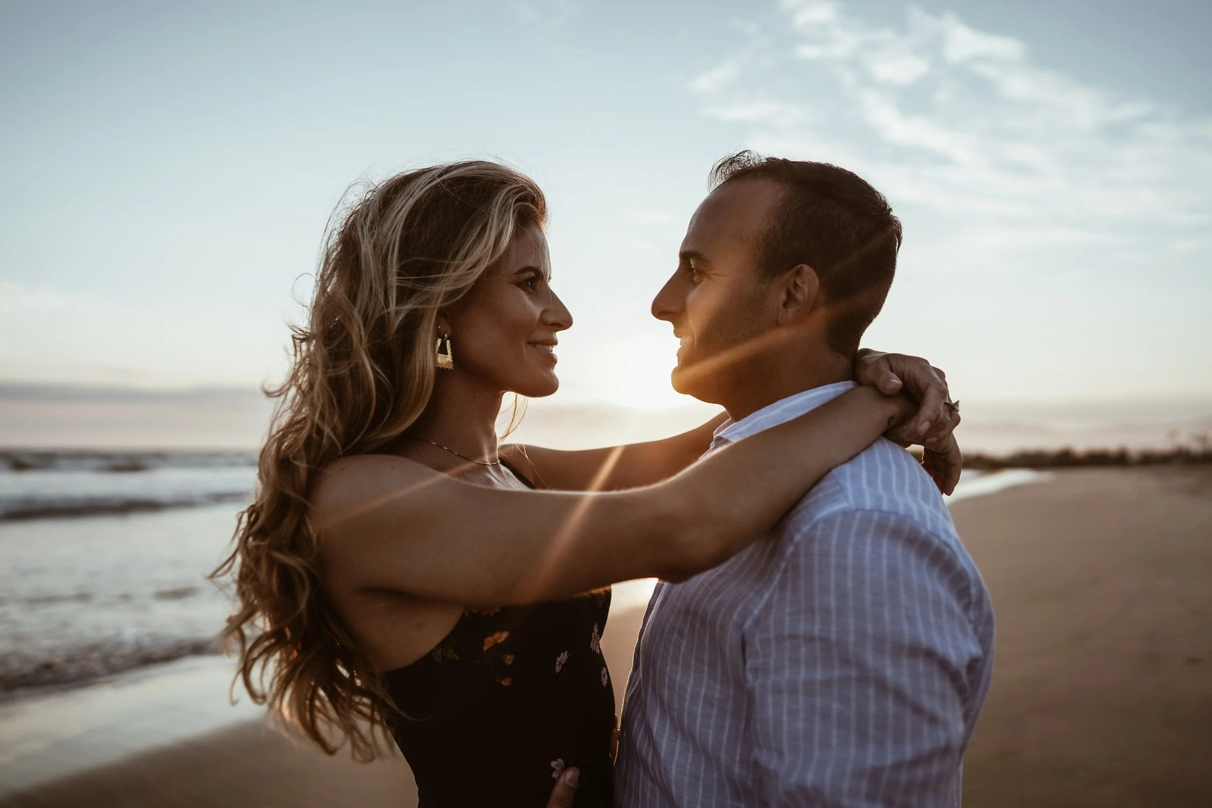 A couple embracing on a beach during sunset, gazing into each other's eyes with the ocean in the background.