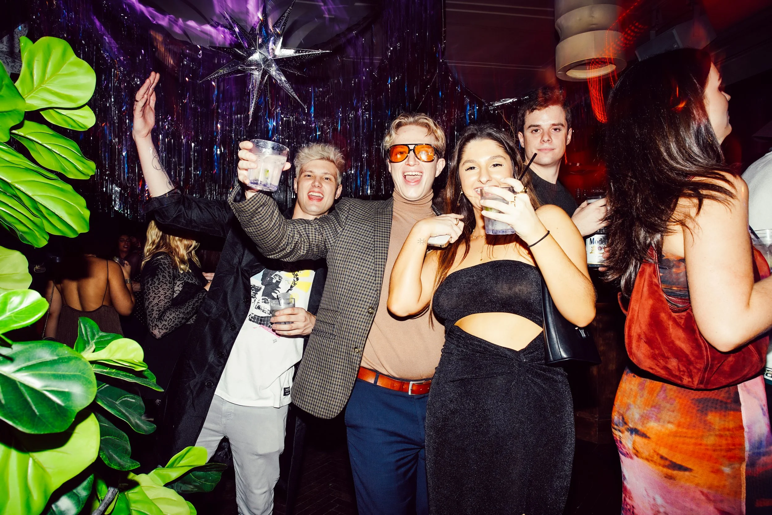 Group of friends celebrating at a party with drinks, colorful decor, and metallic hanging ornaments.