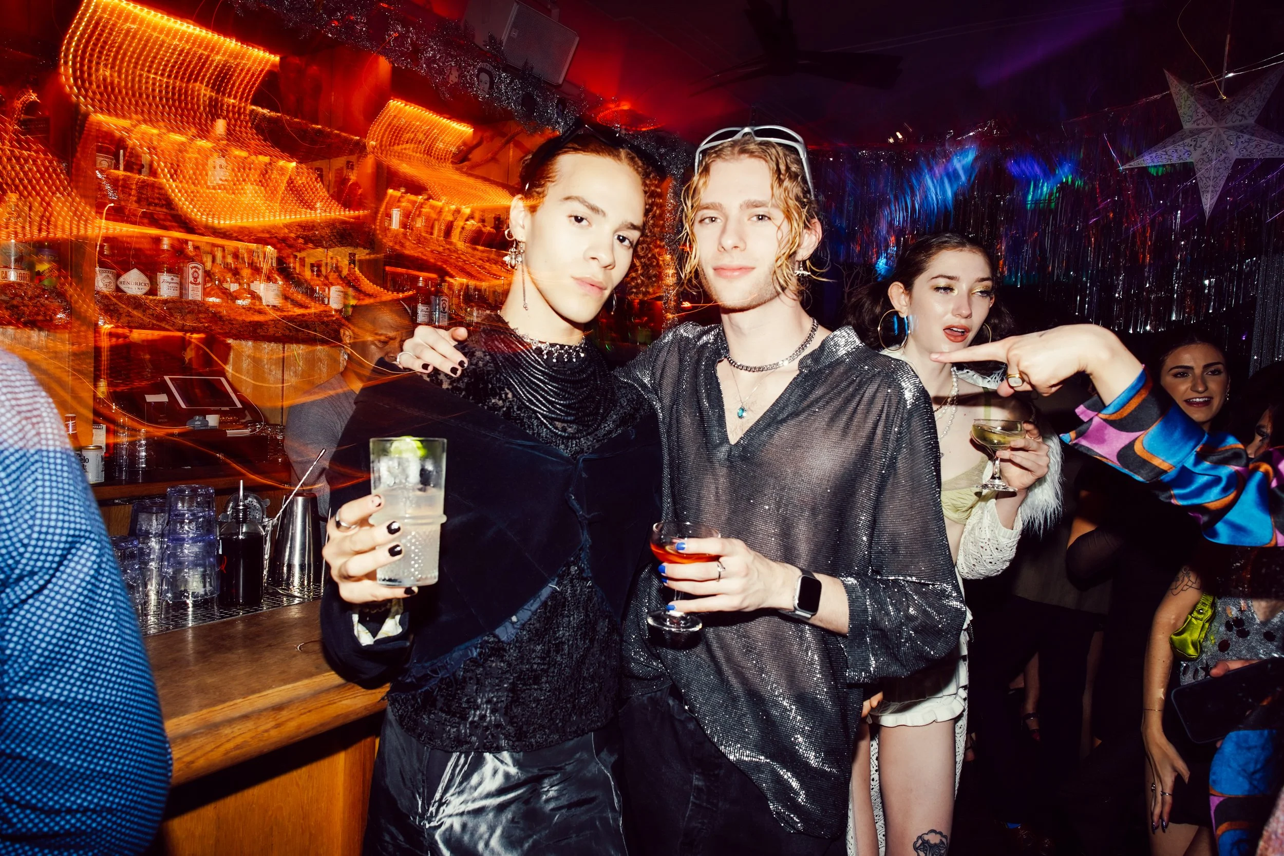 Group of young adults at a party, holding drinks, in a colorful, lively setting with neon lights and decorations.