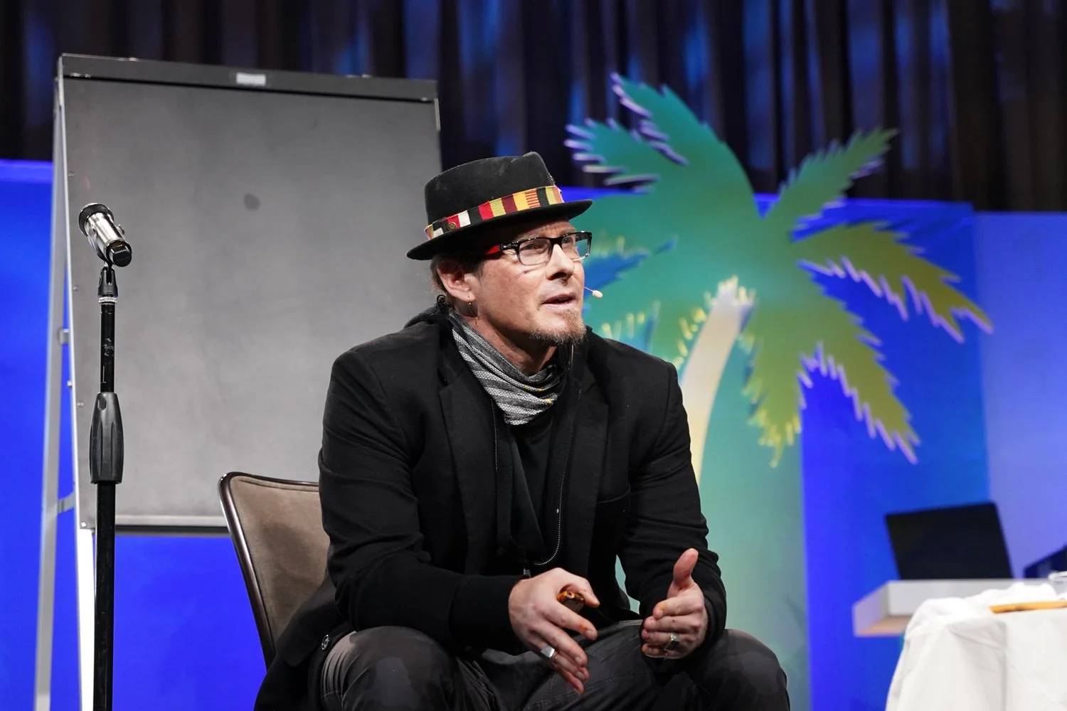 A man sitting on a chair on a stage, wearing glasses, a black hat with a colorful band, a black jacket, and a scarf. There is a microphone stand and a blank whiteboard behind him, with a palm tree graphic in the background.
