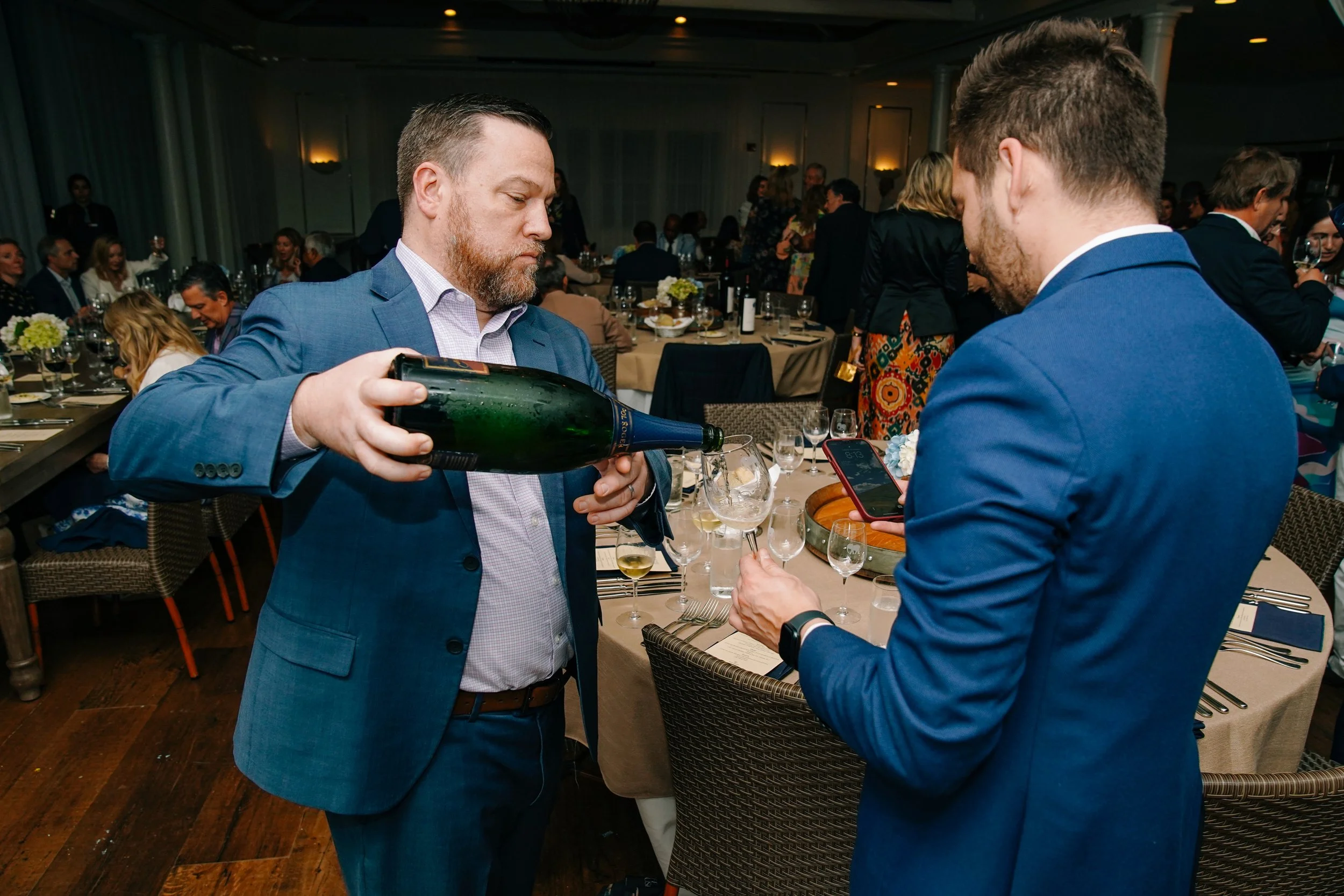 A man in a blue suit pours champagne into a glass at a formal event or banquet, with other guests seated at round tables in the background.