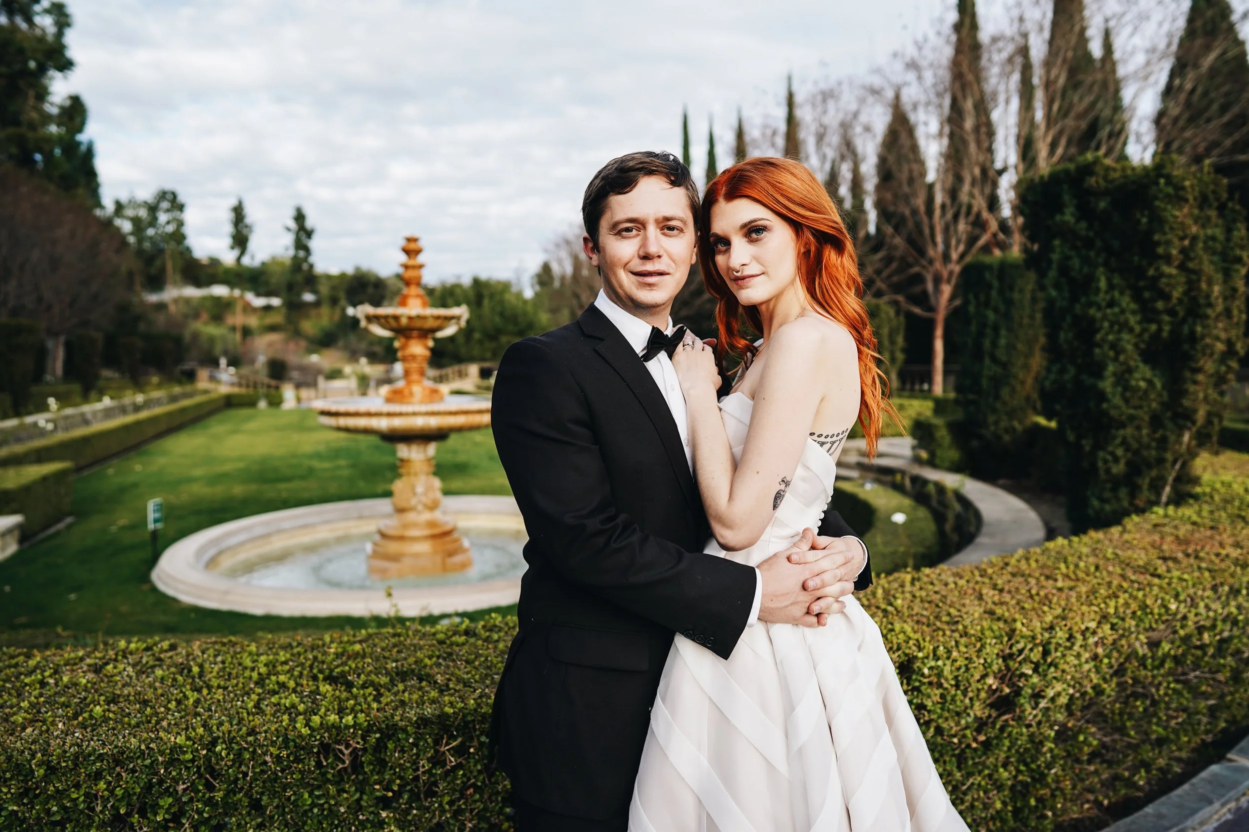 A couple dressed in formal attire, standing out front of a fountain in a garden during daytime, with the man in a tuxedo and the woman in a strapless white dress.
