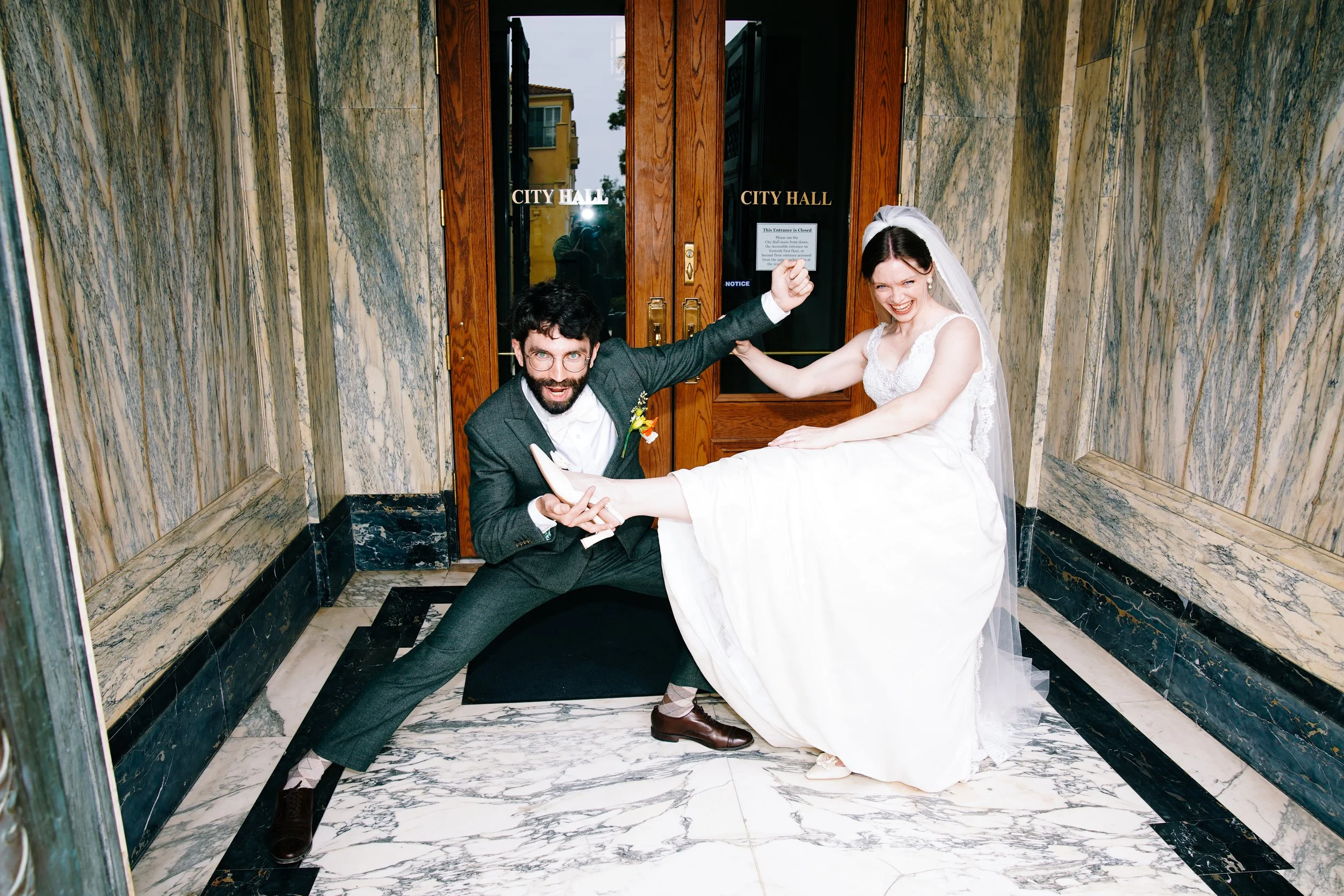 A man in a suit and a woman in a wedding dress are playfully posing in front of the City Hall entrance, with the man holding the woman's leg and both smiling.