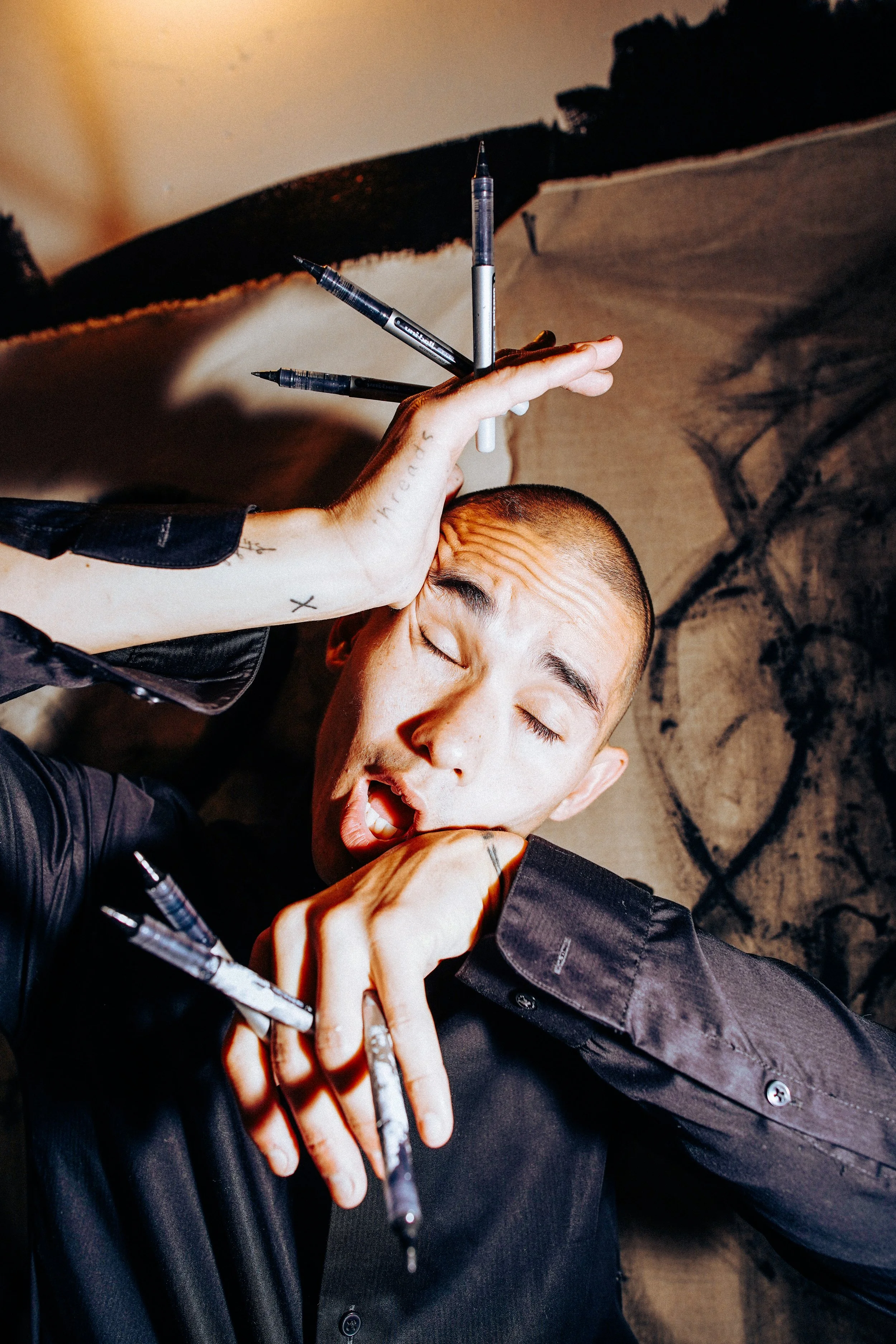 A young man with a shaved head and tattoos, eyes closed, holding multiple black and silver pens against his head, with a surprised or expressive facial expression. Behind him is abstract black and white artwork.