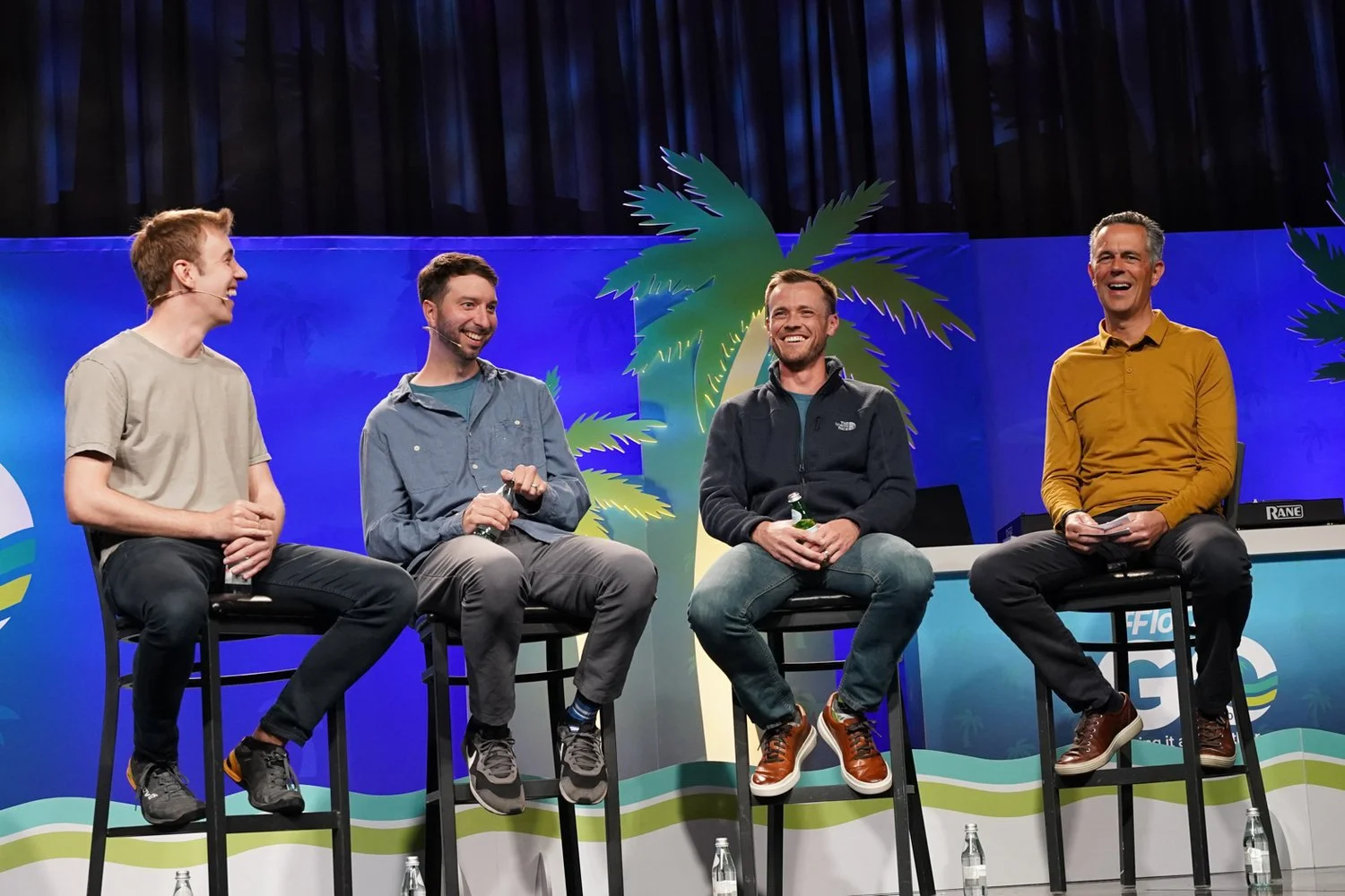 Four men sitting on high chairs on stage, laughing during a panel discussion with a colorful backdrop featuring palm trees and blue skies.