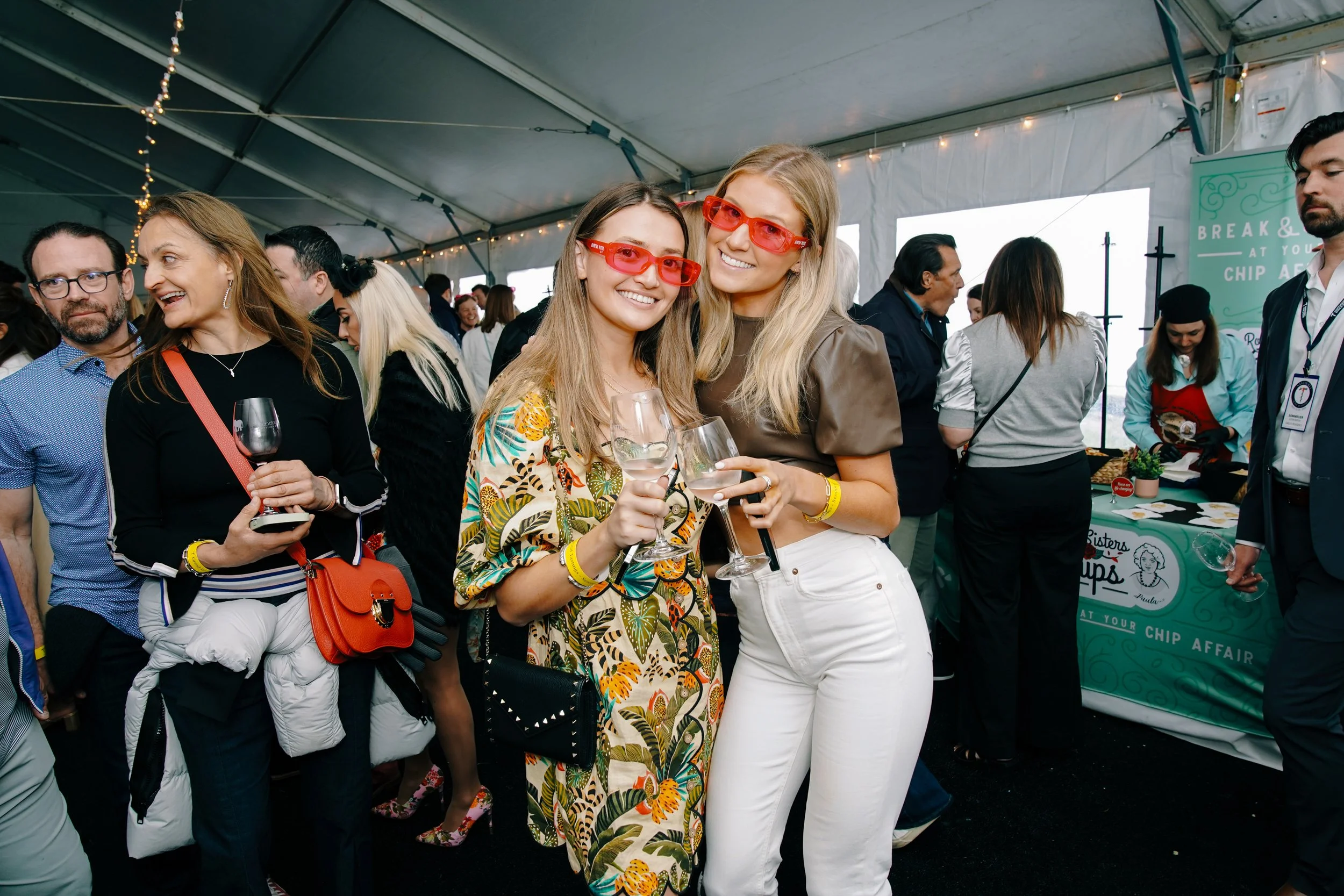 Two young women with blonde hair wearing red glasses smiling and holding wine glasses at a social event inside a tent.