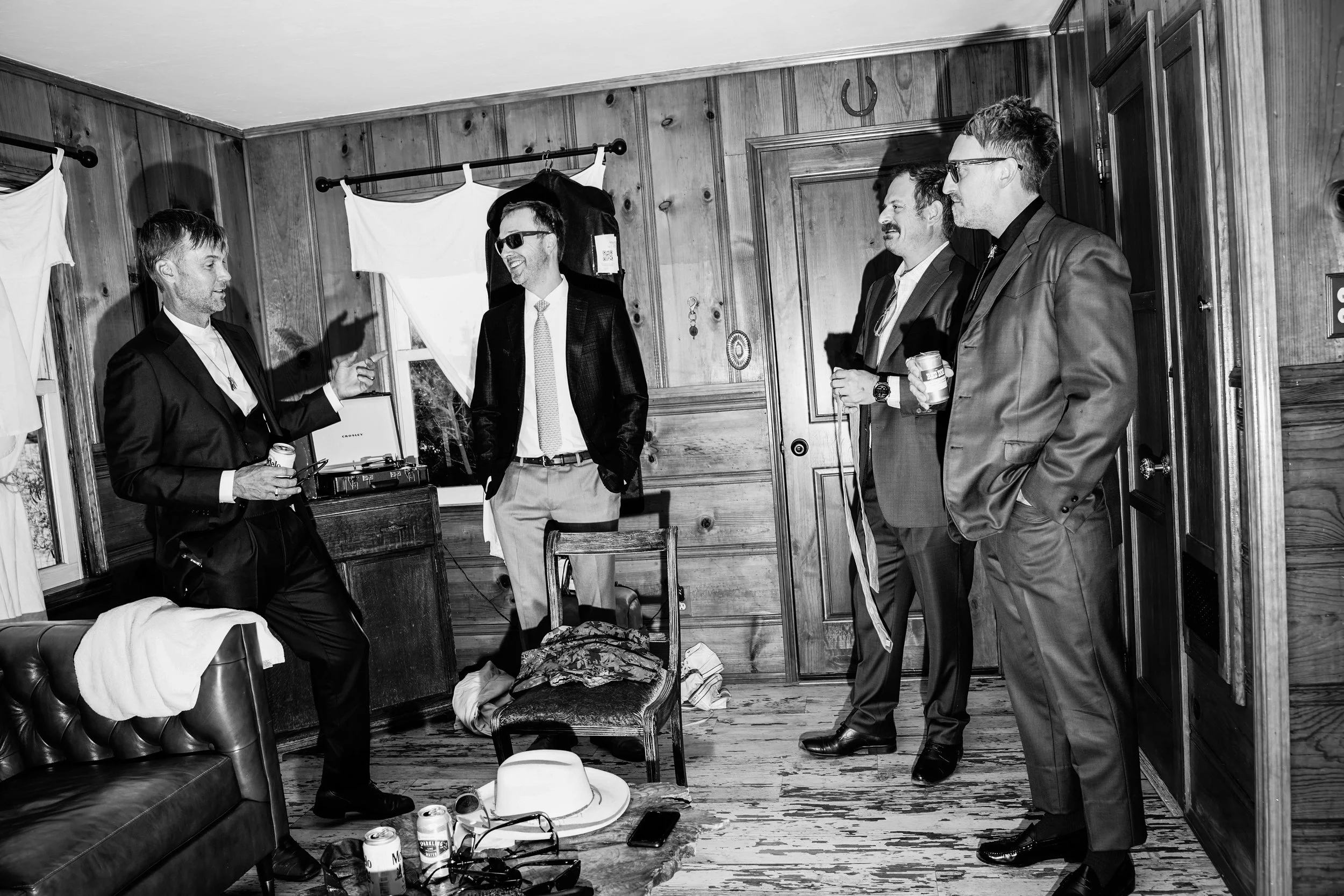 Four men in suits socializing in a wood-paneled room, some holding drinks, with a chair, a hat on the floor, and window with curtains in the background.