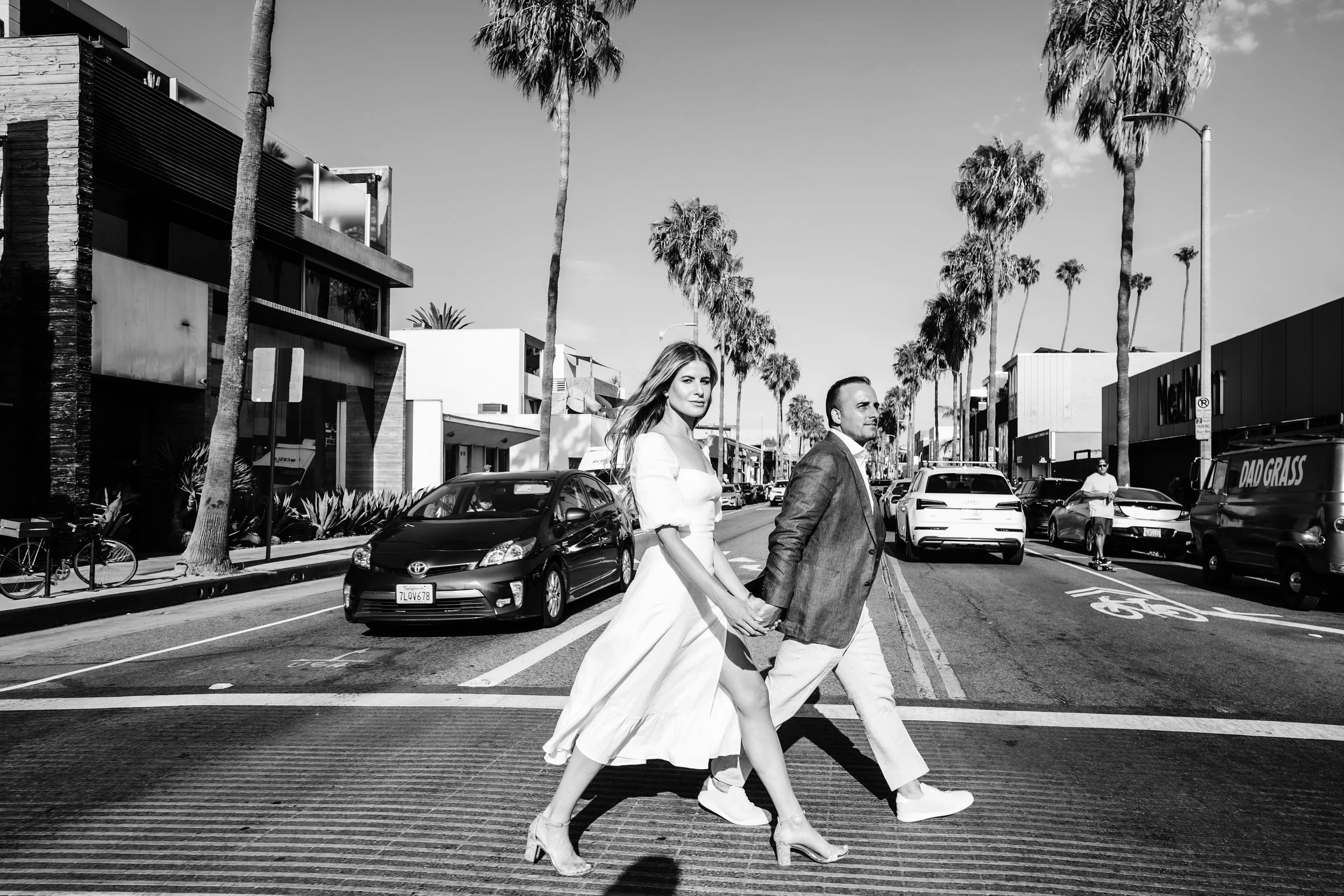 A couple walking hand in hand across a city street, with palm trees and modern buildings in the background, black and white photo.