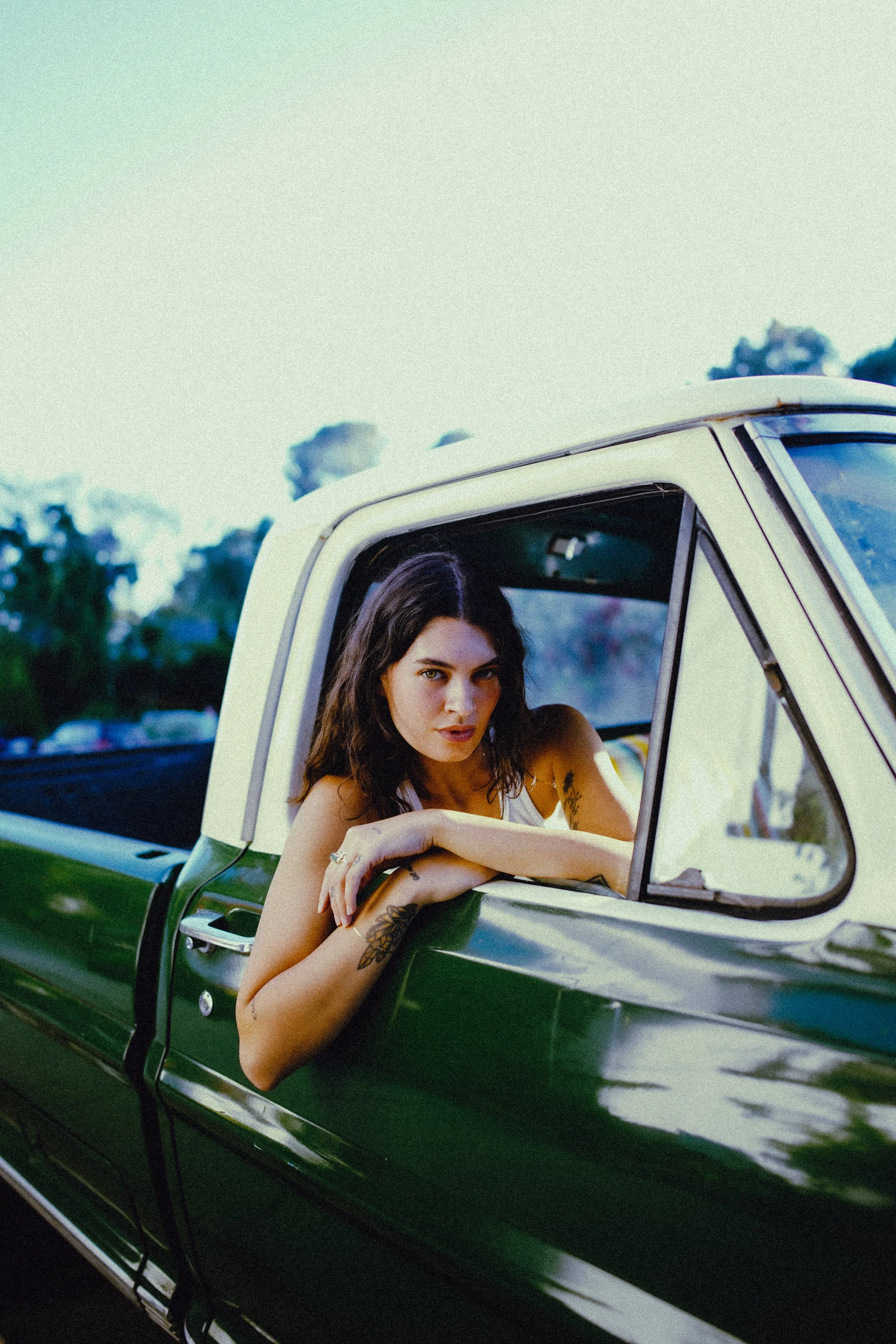 A young woman with dark wavy hair leaning out of the passenger window of a vintage green and white pickup truck, looking at the camera.