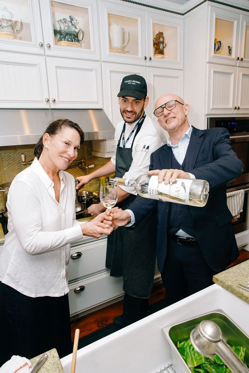 A woman is receiving a glass of white wine from a man in a suit, while a chef behind them pours more wine into the glass, in a kitchen setting.