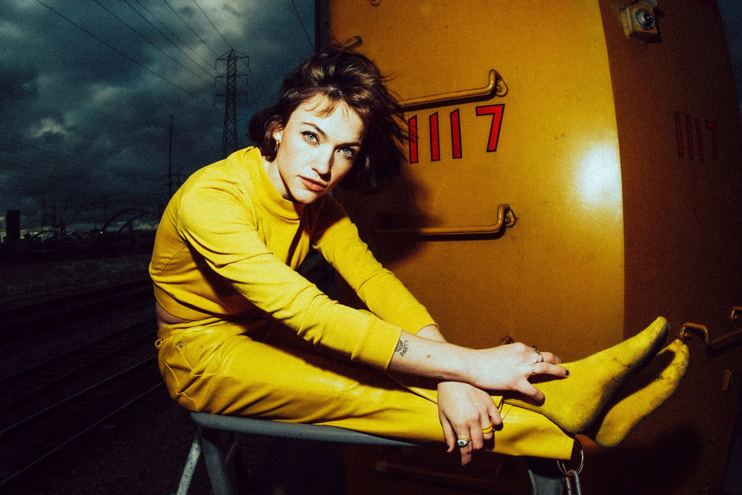 A woman in a yellow outfit sitting on a bench, outdoors at dusk, with dark clouds and power lines in the background.