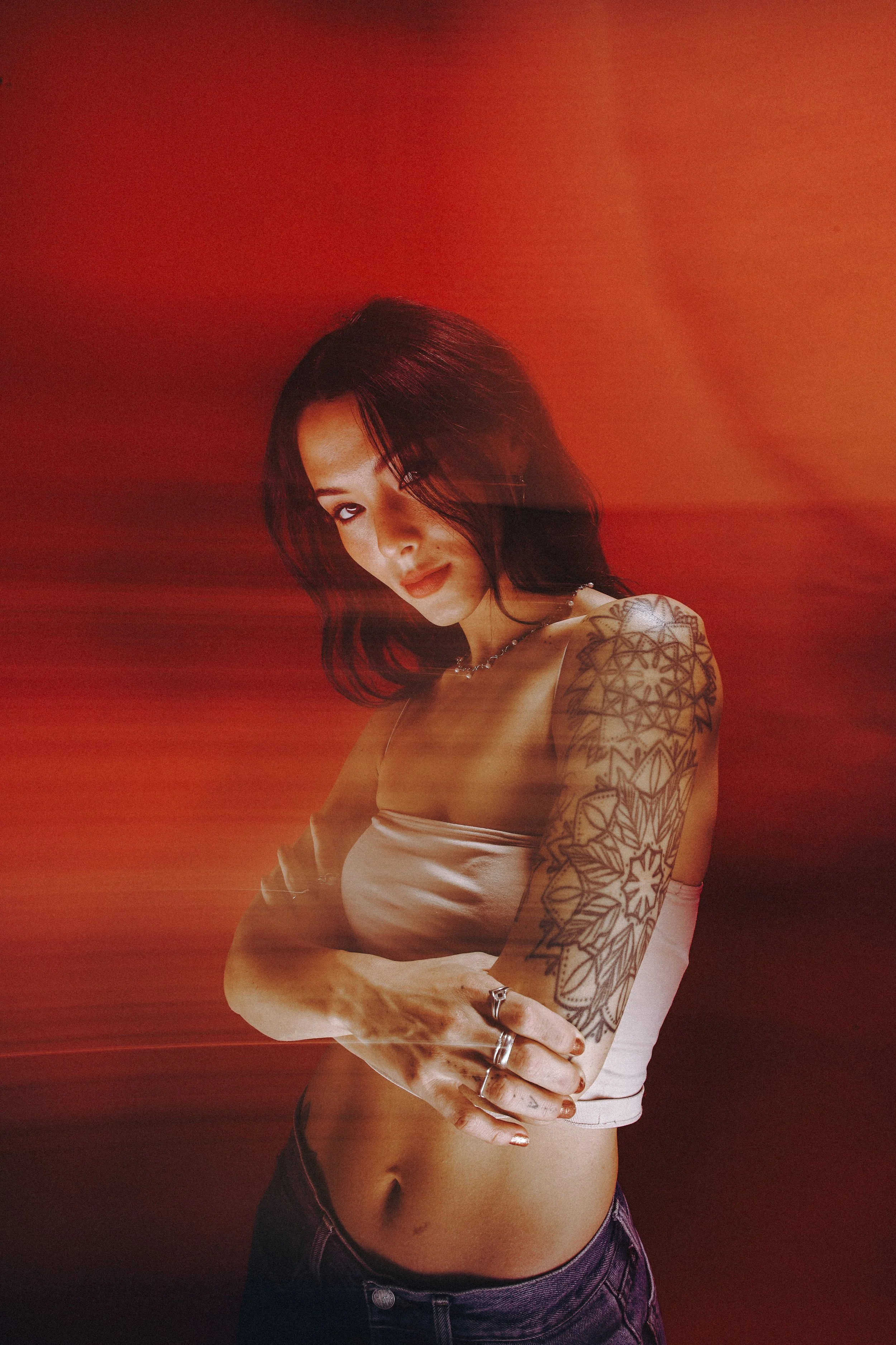 A woman with dark hair and tattoos on her arm, wearing a beige tube top and dark pants, stands against a red background with motion blur effects.