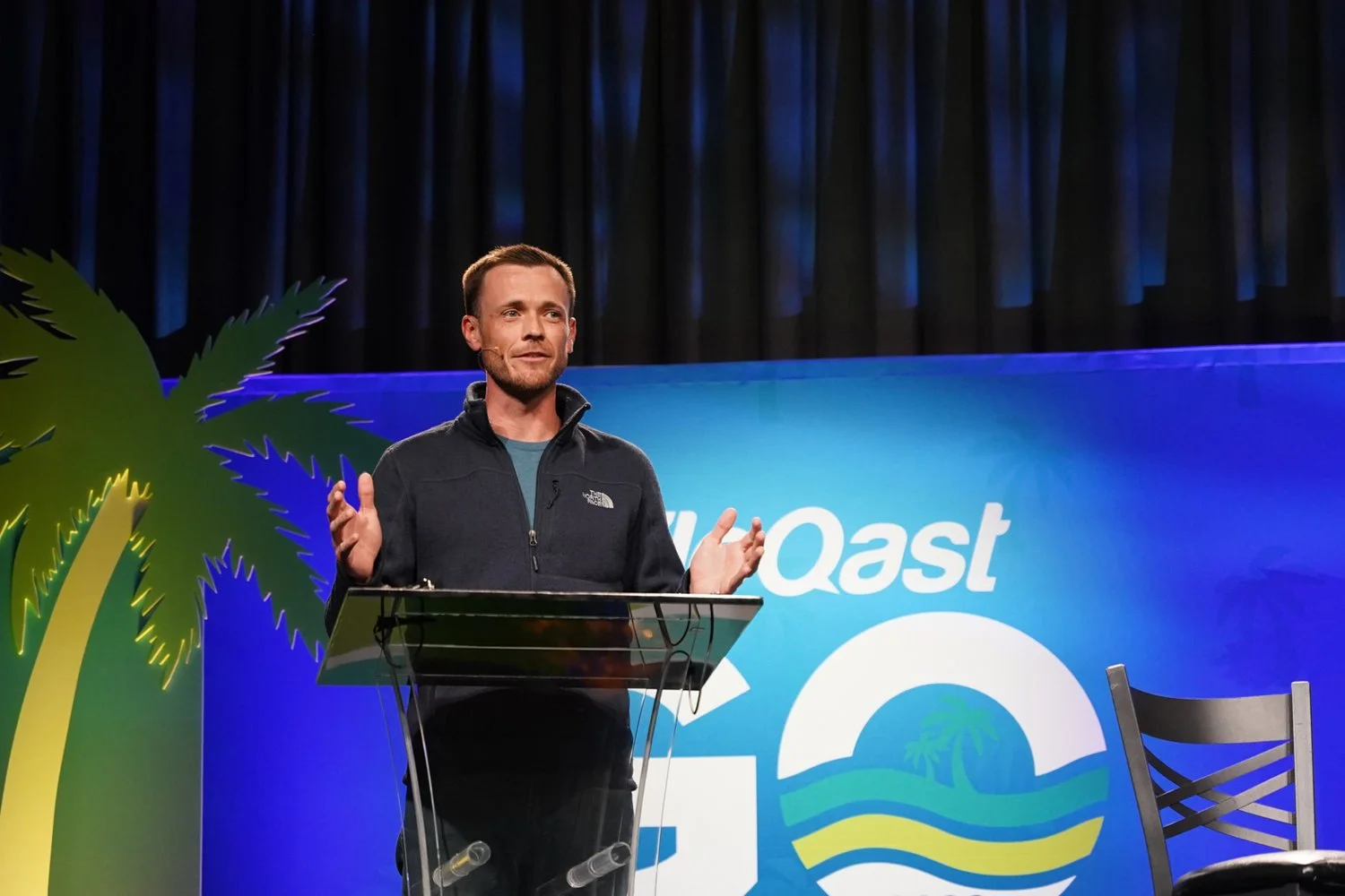 A man speaking at a podium with a backdrop displaying the words 'WQast' and a logo of a sunset over water and palm trees, with a large fake palm tree on the left side of the stage.
