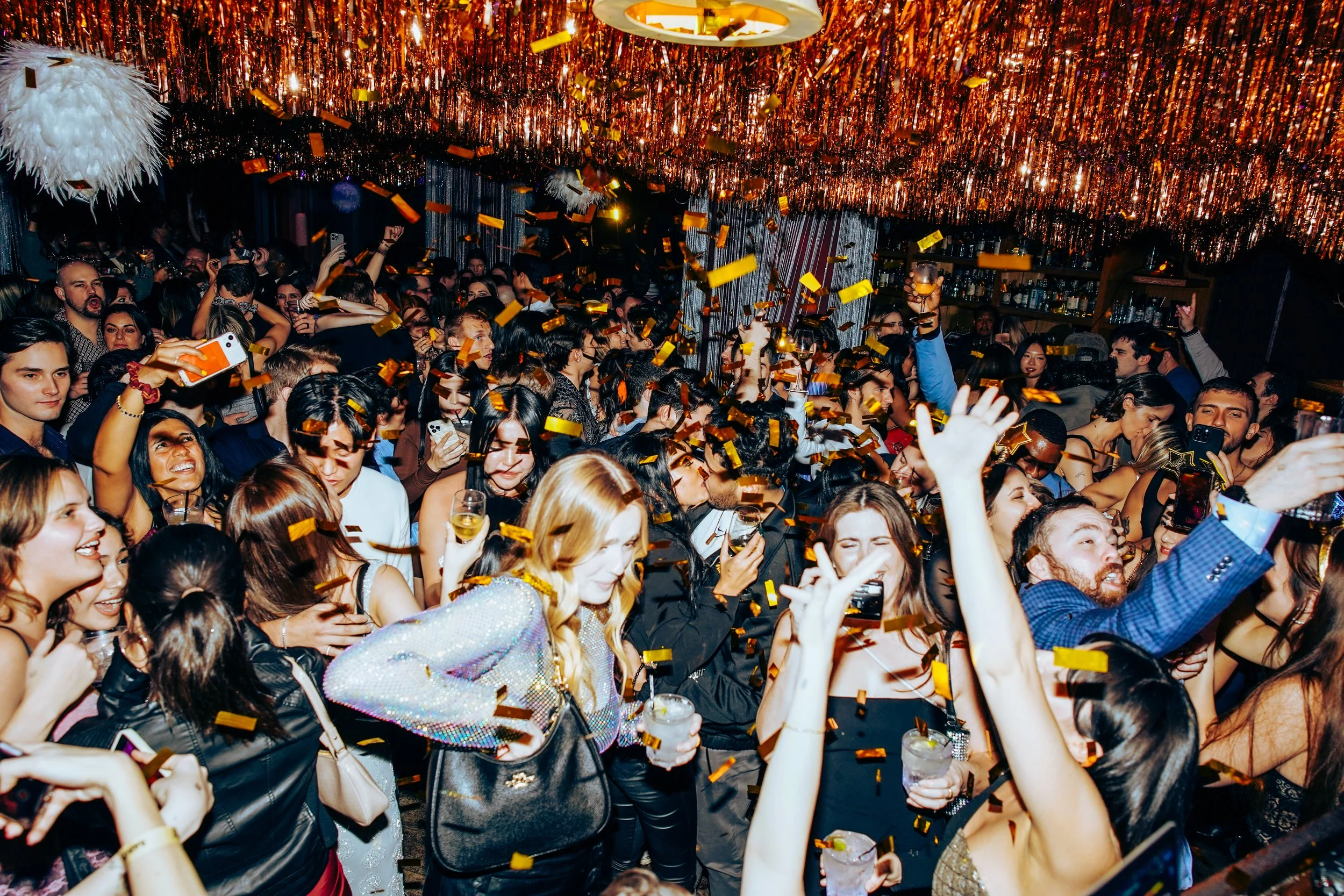 Crowd of people celebrating at a party with gold and black confetti falling and shiny decorations on the ceiling.