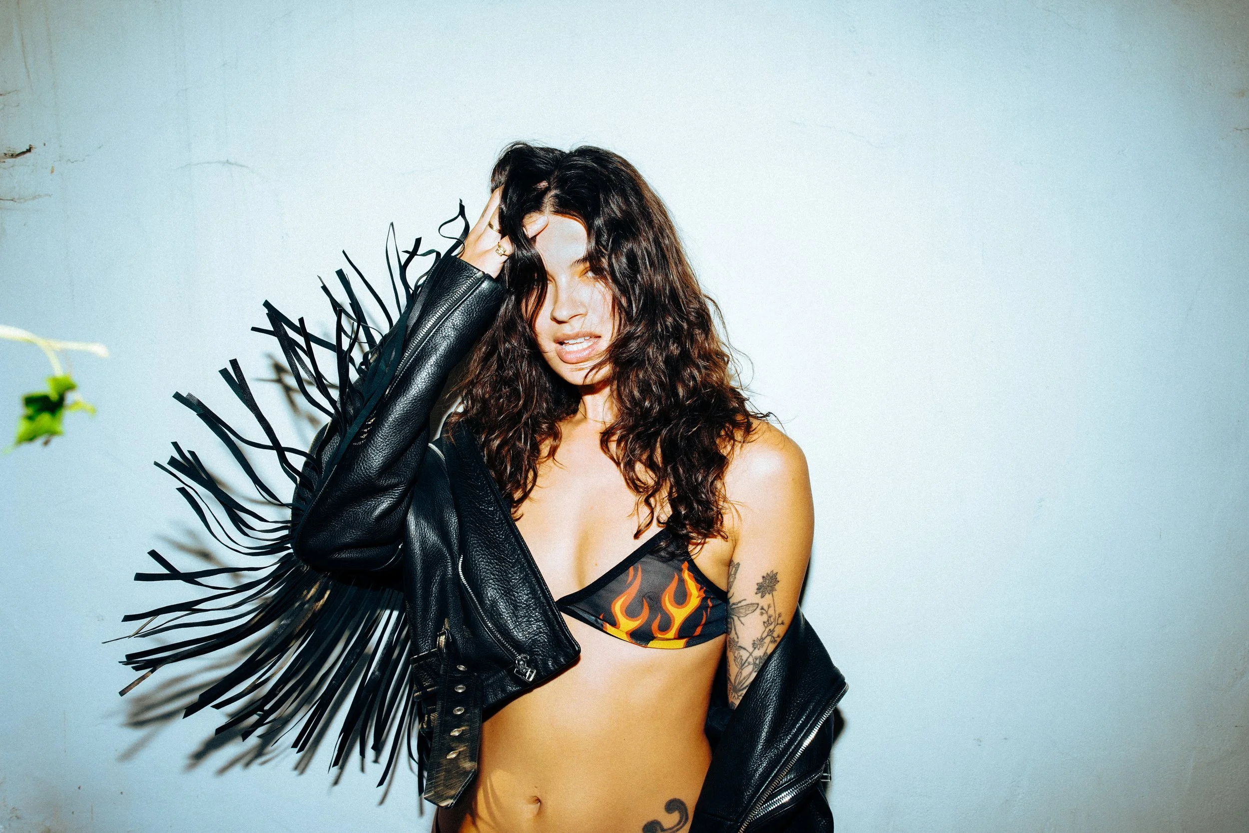 A woman with curly brown hair wearing a flame-patterned bikini top, a black leather jacket with fringe, and tattoos, standing against a plain wall.