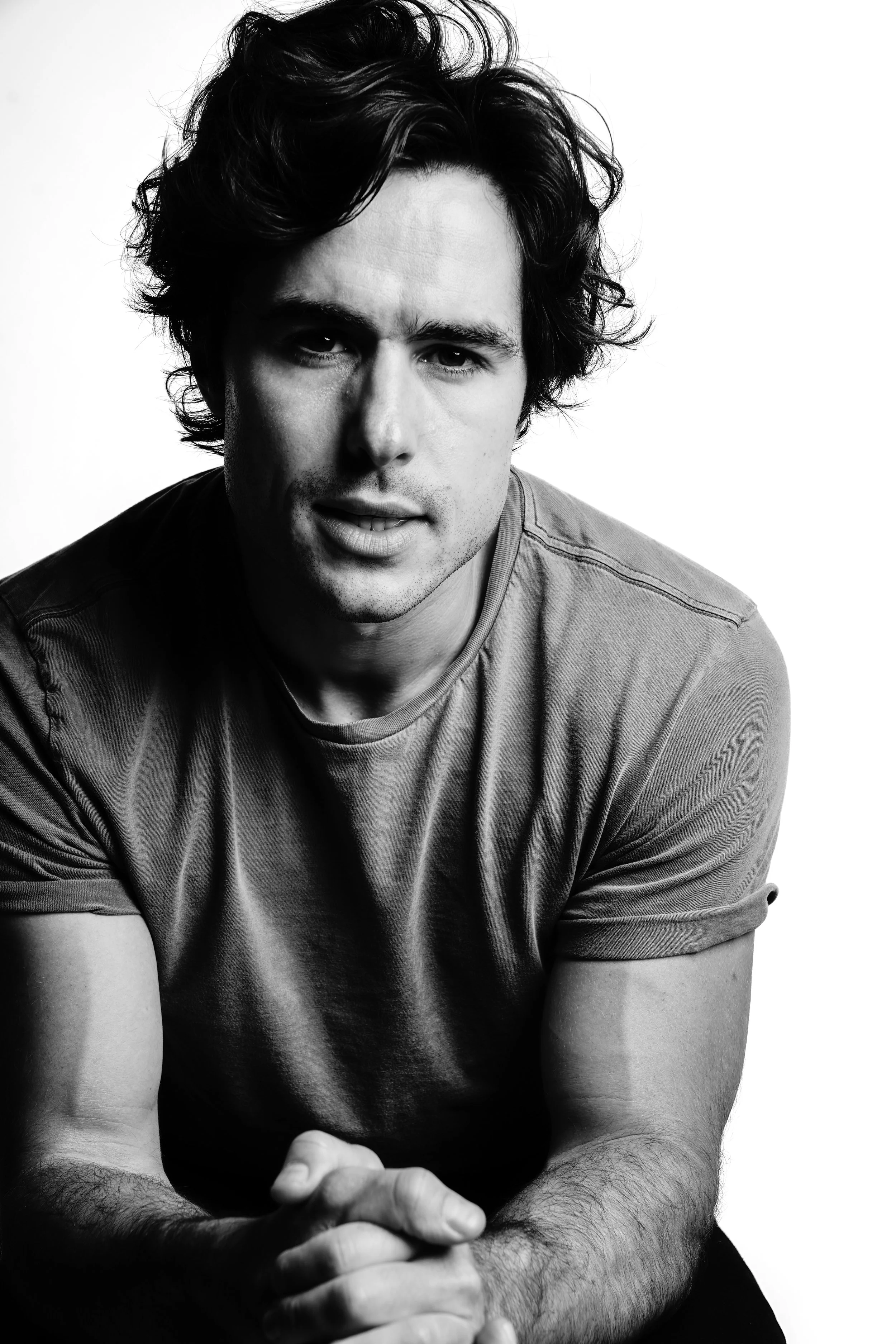 A black and white portrait of a young man with tousled hair, wearing a T-shirt, looking directly at the camera with a slight smile, with a plain background.