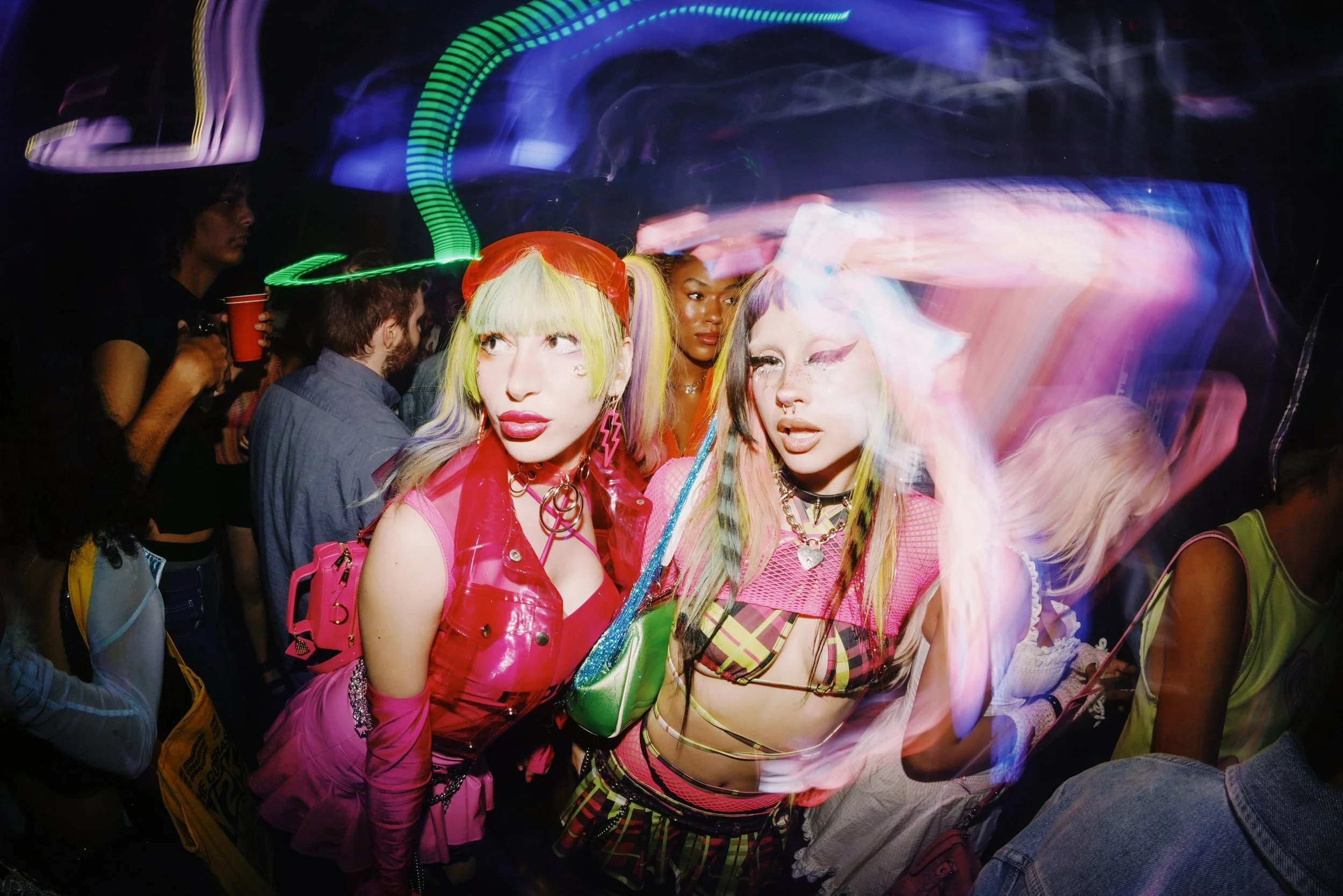 Two women with colorful hair and bold fashion poses in a crowded, vibrant nightclub with light streaks.