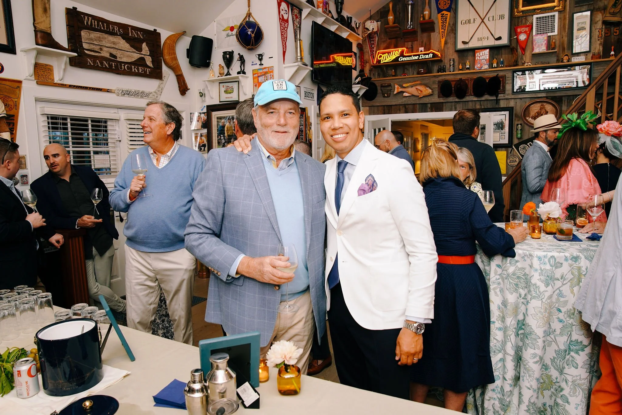 People socializing at a party or gathering in a decorated room with nautical and vintage decor, including a fish mounted on the wall, flags, and signs. Two men are in the foreground, one older with a blue cap and light gray blazer, the other younger 
