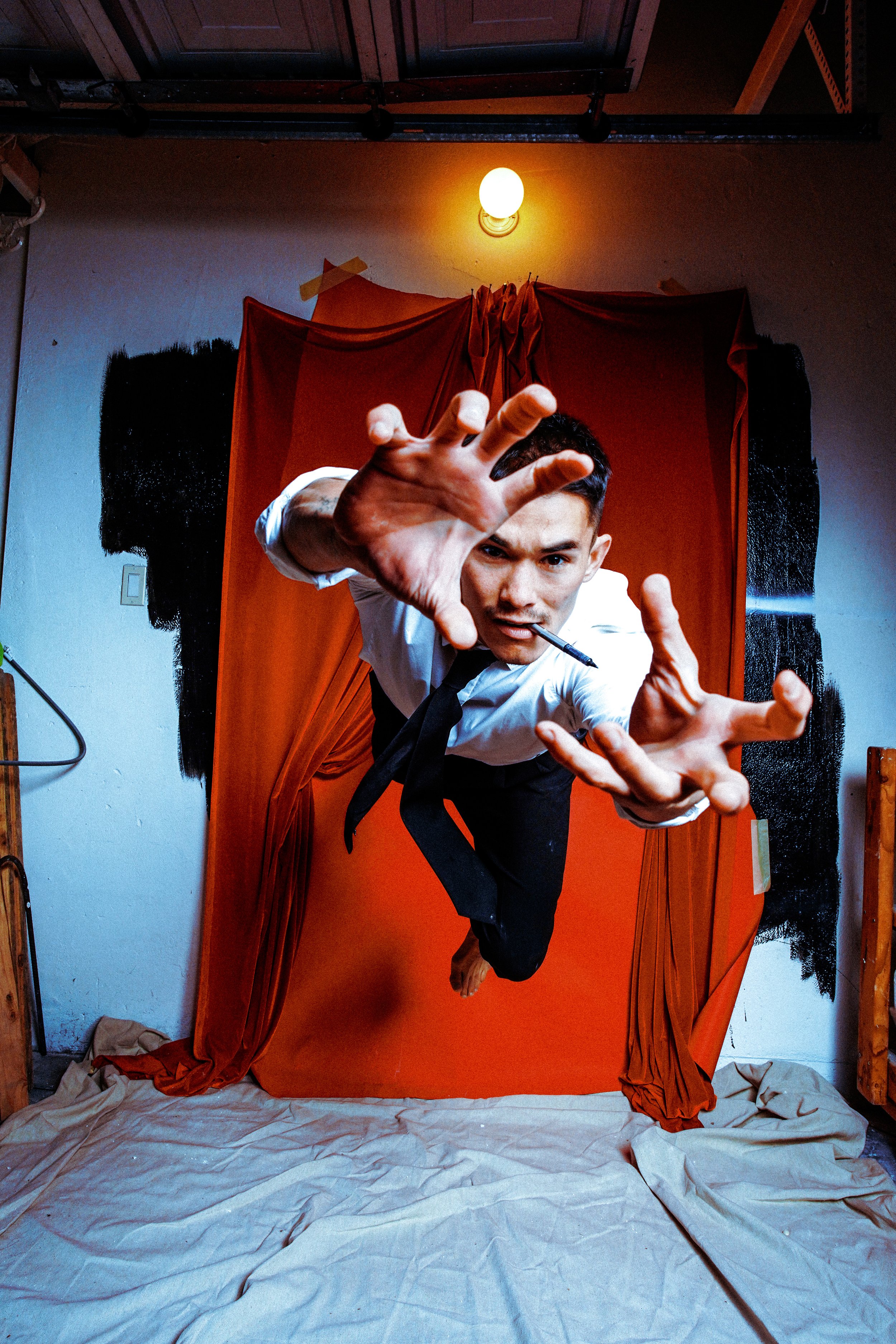 A man in formal attire, holding a pen in his mouth, is reaching toward the camera while appearing to float or jump in front of an orange backdrop with black paint on the sides. The scene is inside a room with a white wall, some wooden furniture, and 