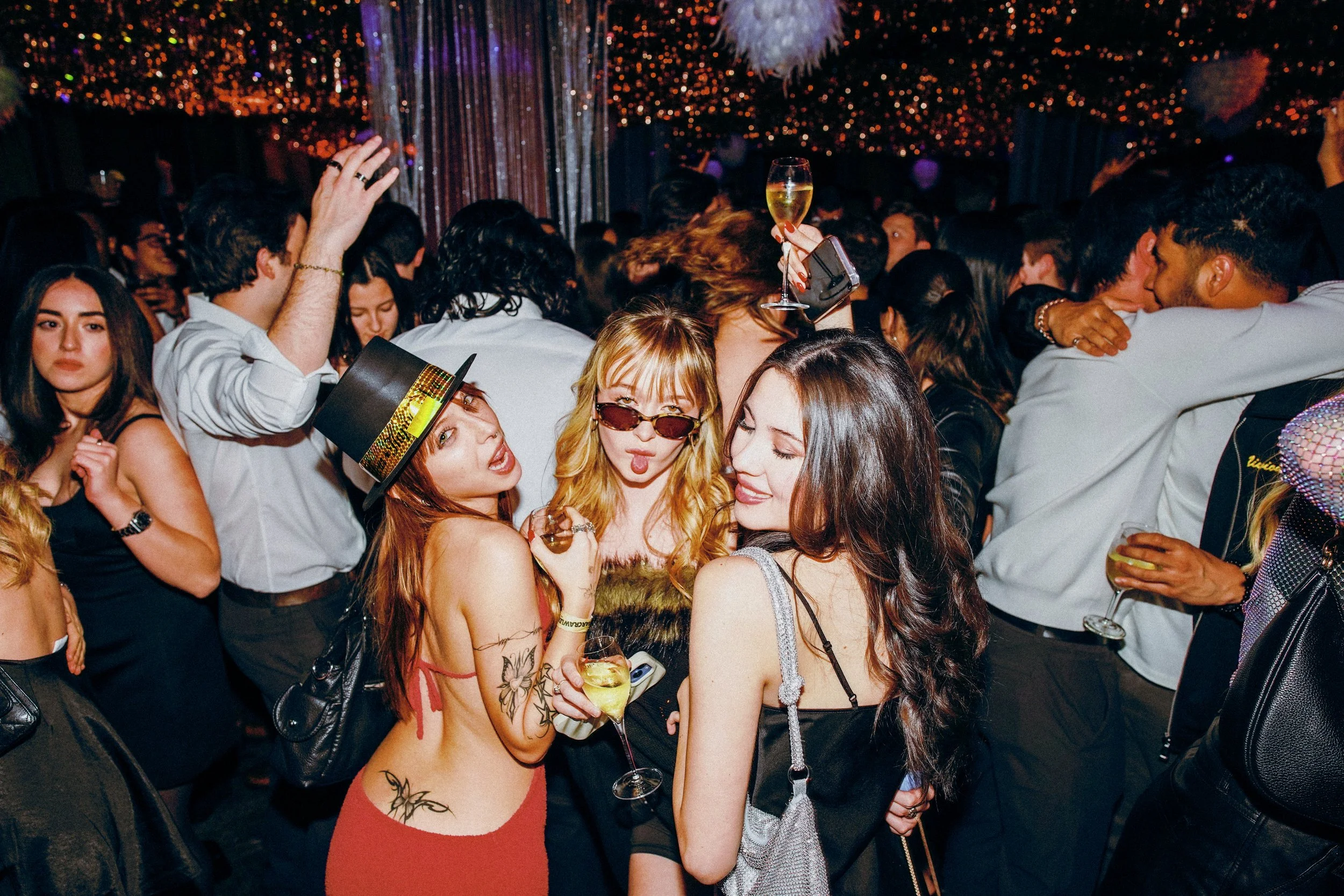 A crowded party scene with people dancing and socializing, featuring three women in the foreground enjoying drinks and posing for the camera, with disco lights and decorations in the background.