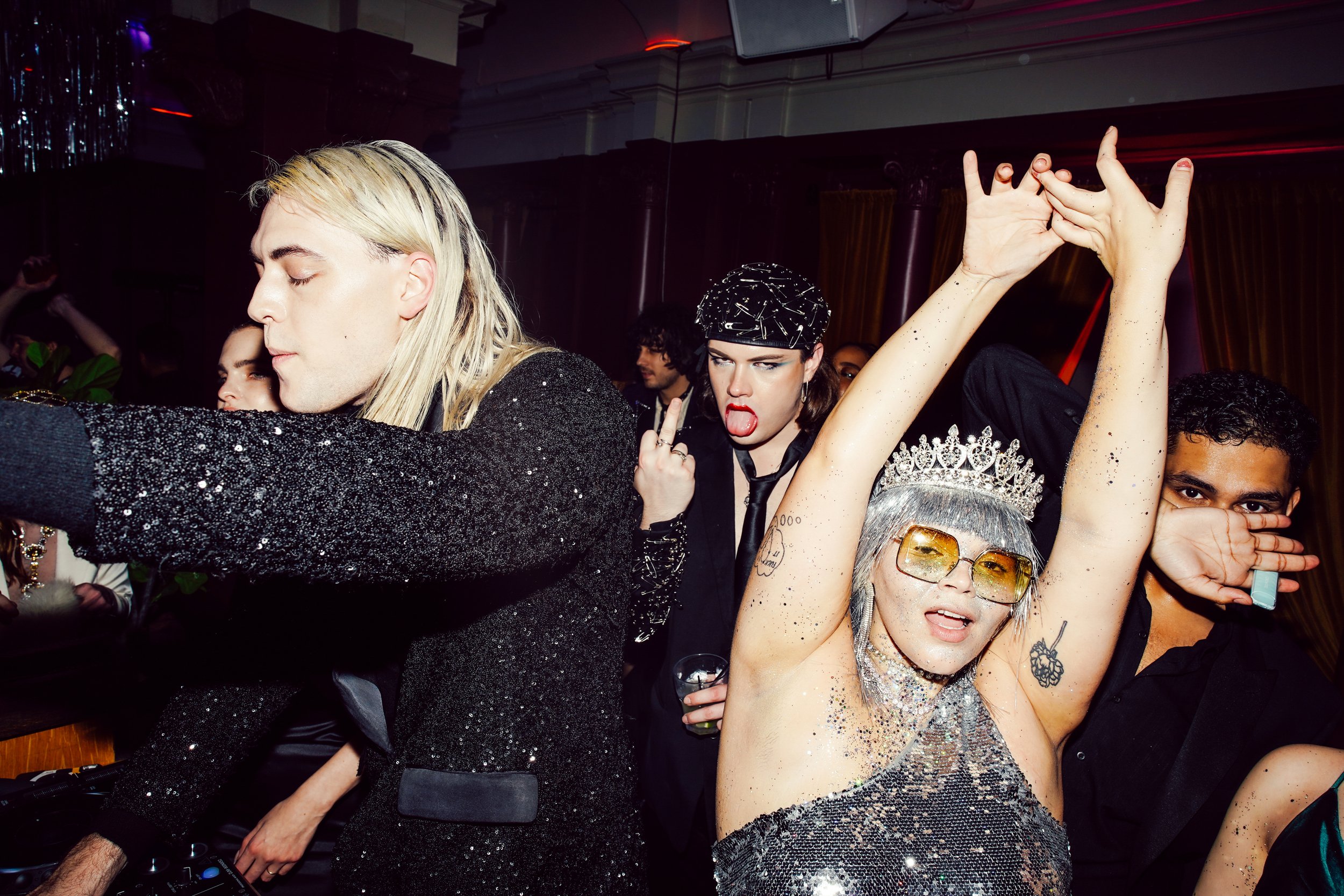 People dancing at a party, with a woman wearing a tiara and sunglasses making a peace sign and a man with long blond hair wearing a glittery black jacket in the foreground. Others are seen in the background enjoying the event.