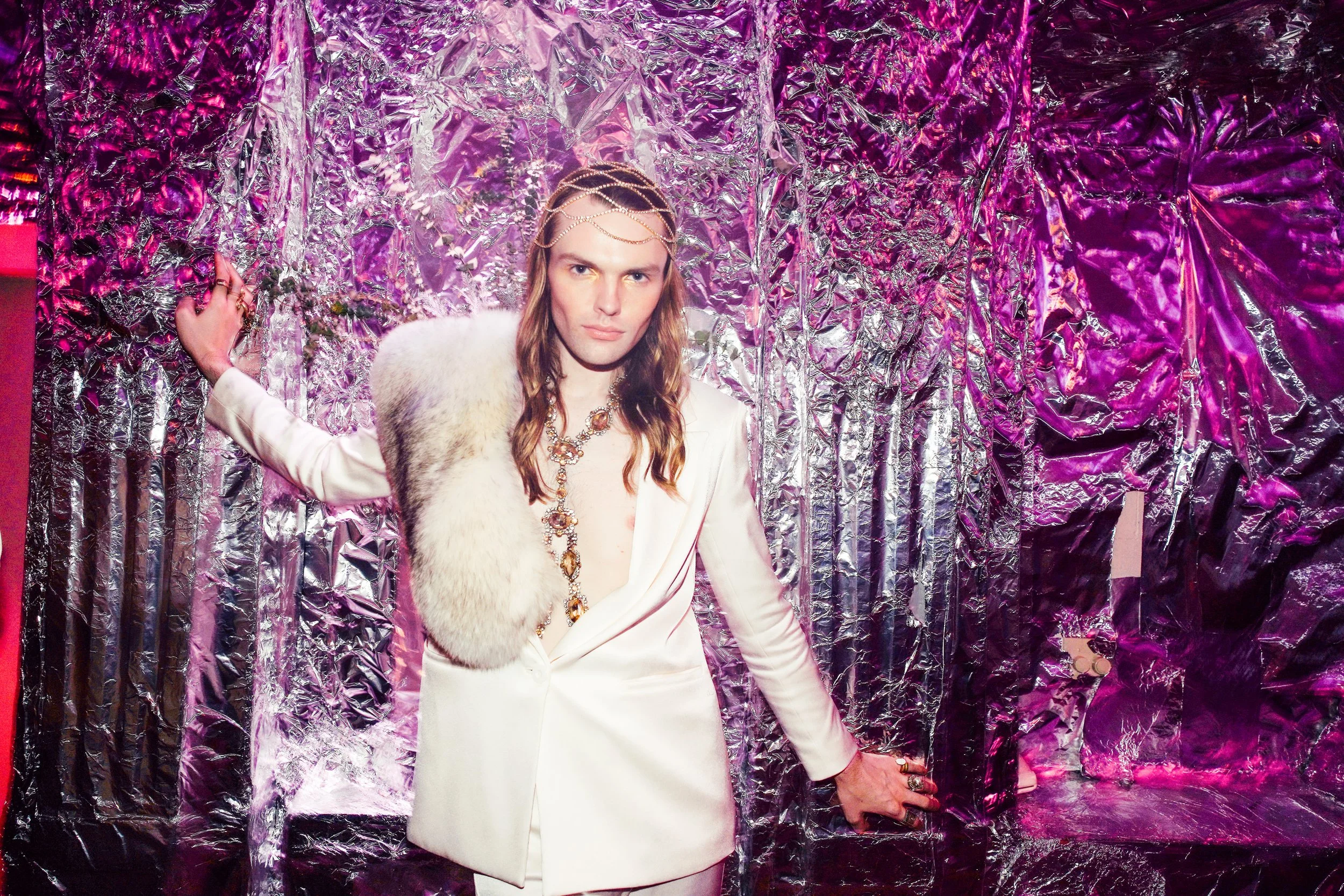 A man with long hair is wearing a white suit with a fur stole and ornate jewelry, standing against a crinkled metallic purple background.