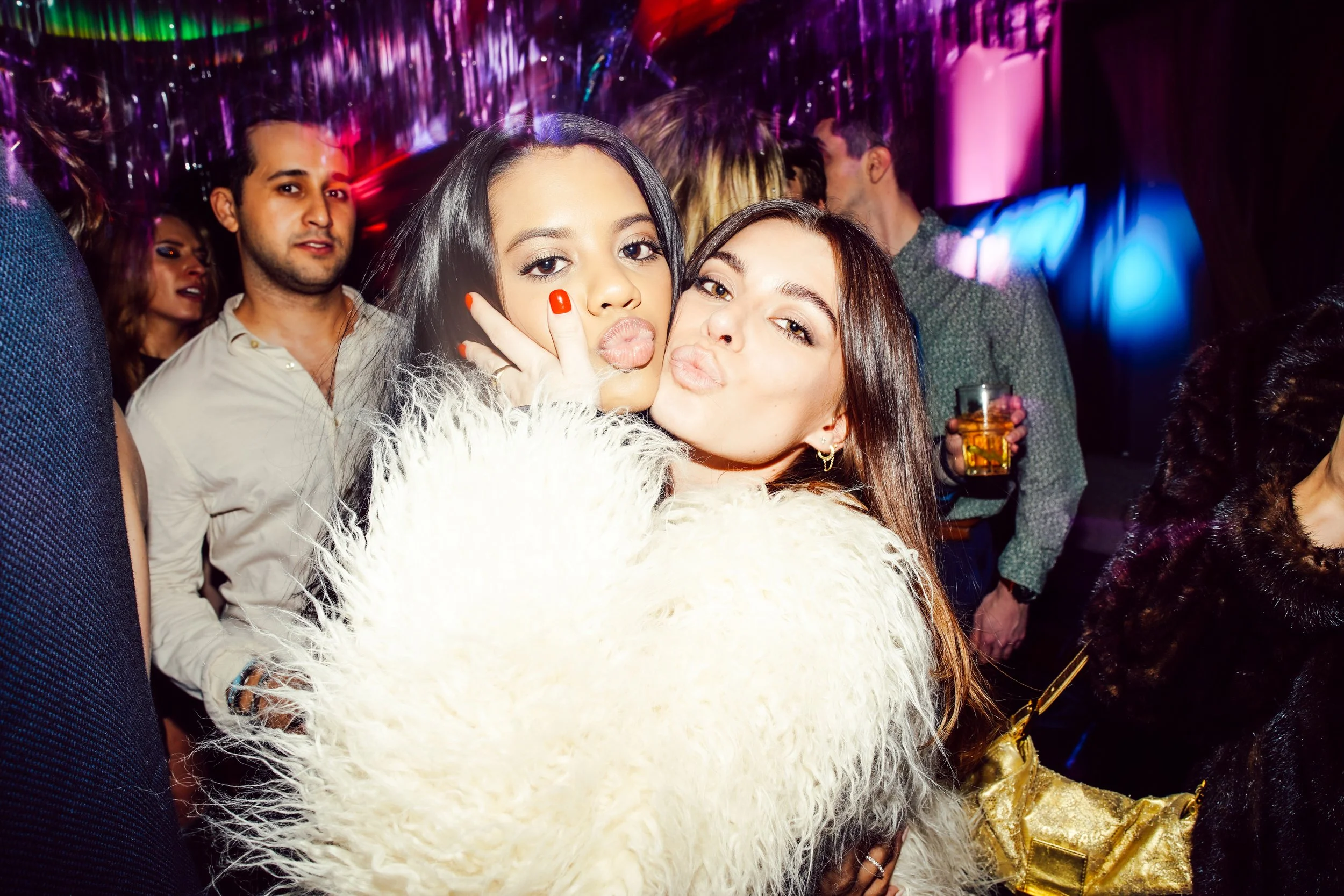 Two women puckering their lips and making a peace sign at a party, with colorful decorations and other party-goers in the background.