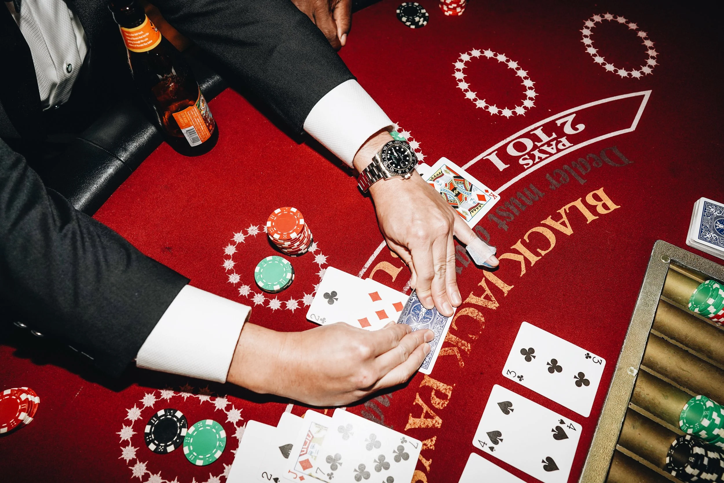 A person in a tuxedo playing blackjack at a casino table, handling playing cards with chips and a beer bottle nearby.