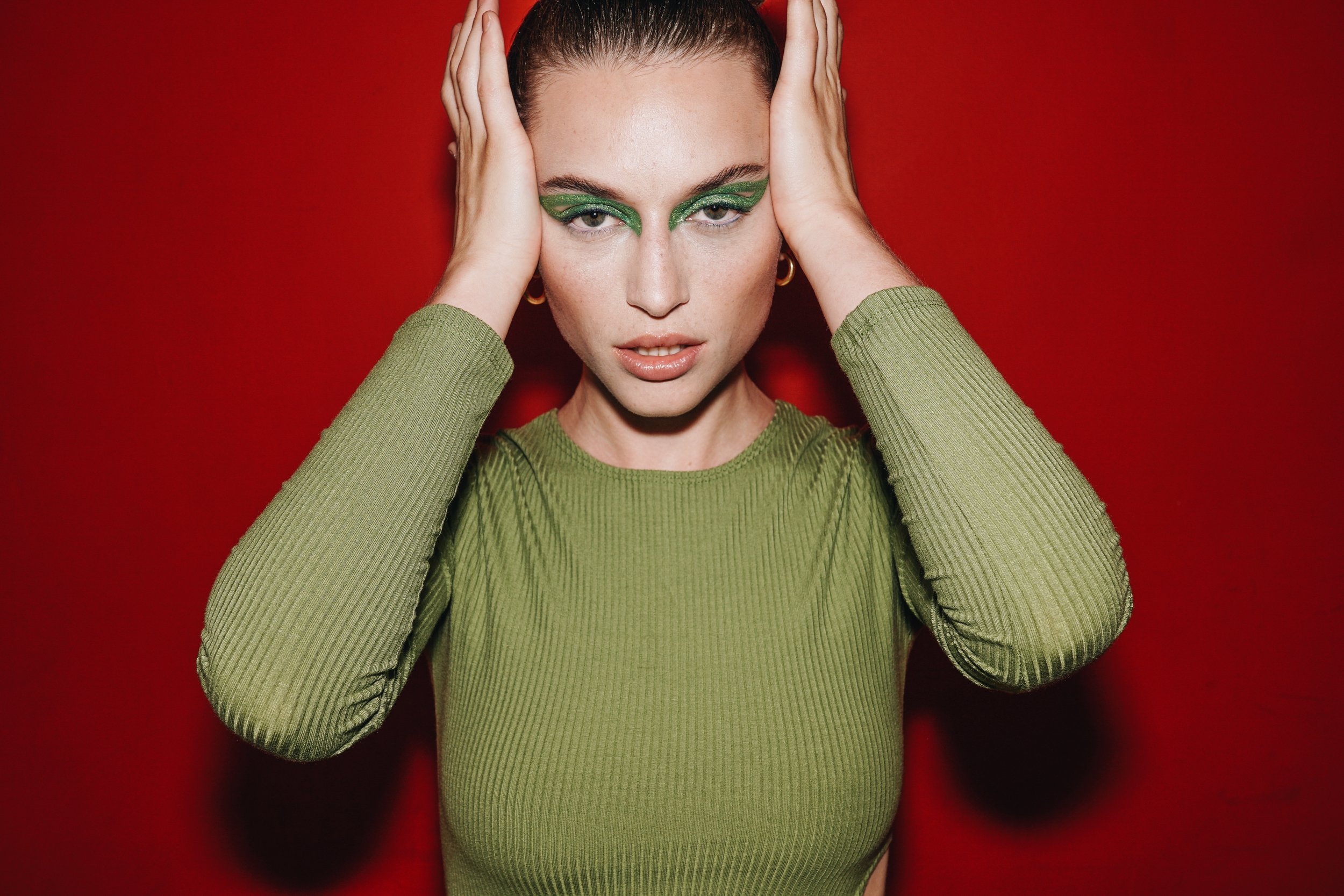 A woman with green eye makeup and a green ribbed top holds her hands to her head against a red background.