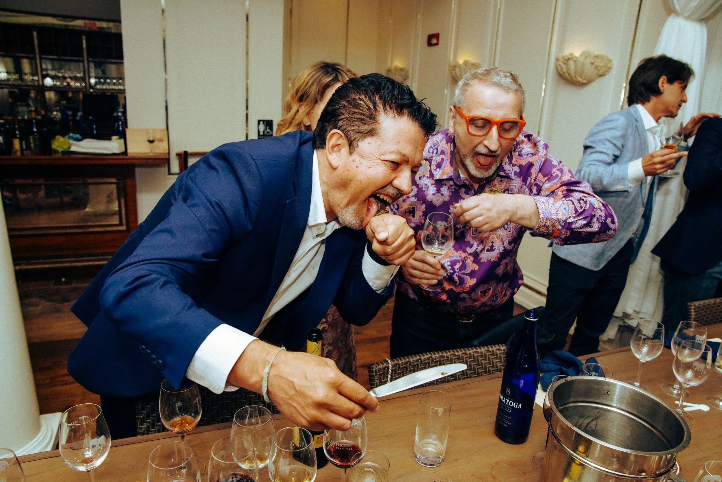 A group of people having fun at a social gathering, with two men in the foreground laughing and pouring drinks at a table, surrounded by wine glasses and a bottle of wine.