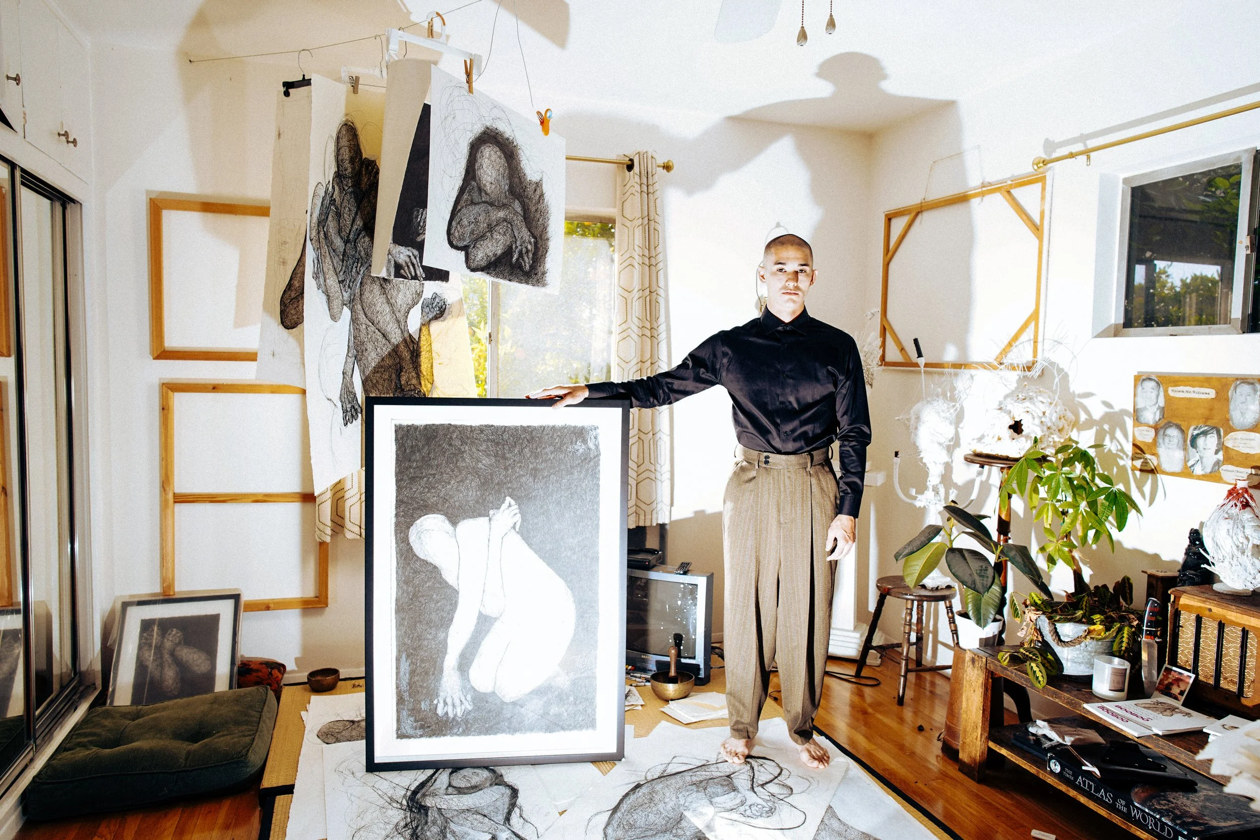 Artist standing barefoot in a cluttered art studio, surrounded by portraits and sketches, with a large framed drawing in front of her, wearing a black blouse and beige pants.
