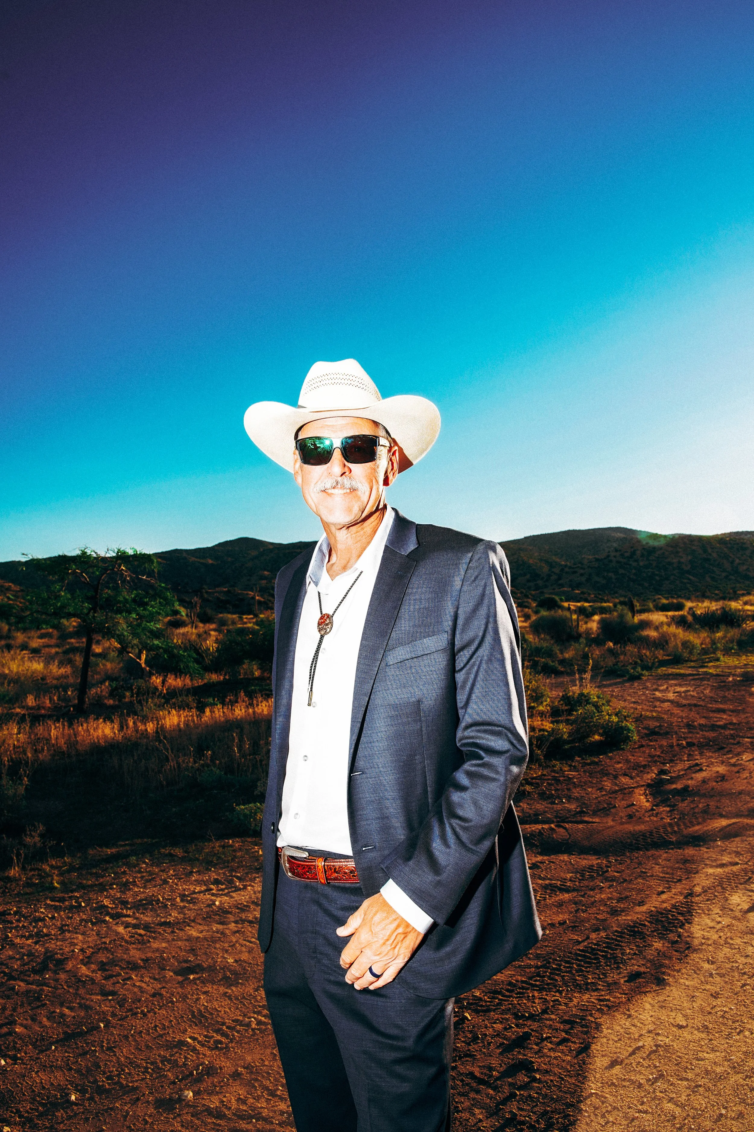 A man in a suit with sunglasses and a cowboy hat standing outdoors in a desert landscape with mountains in the background.