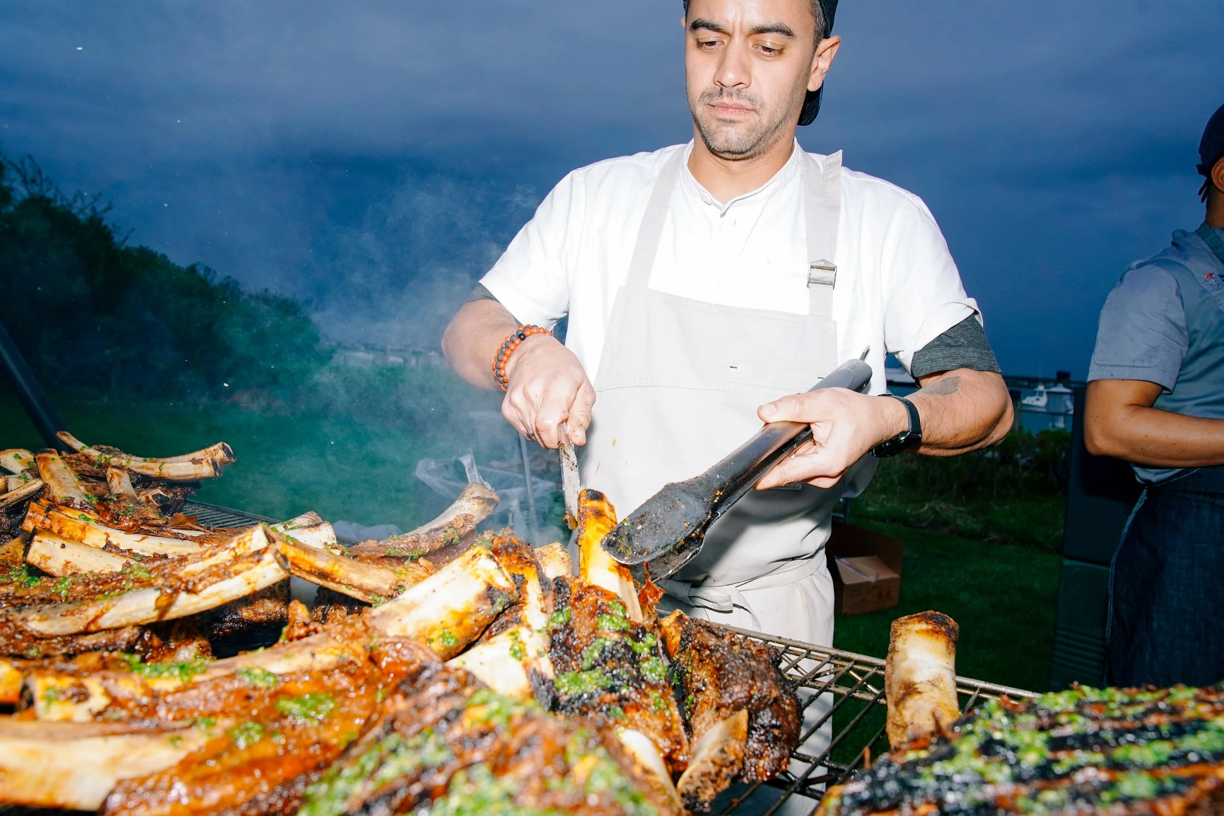 A man wearing a white apron cooks lamb chops on a grill outdoors during dusk.